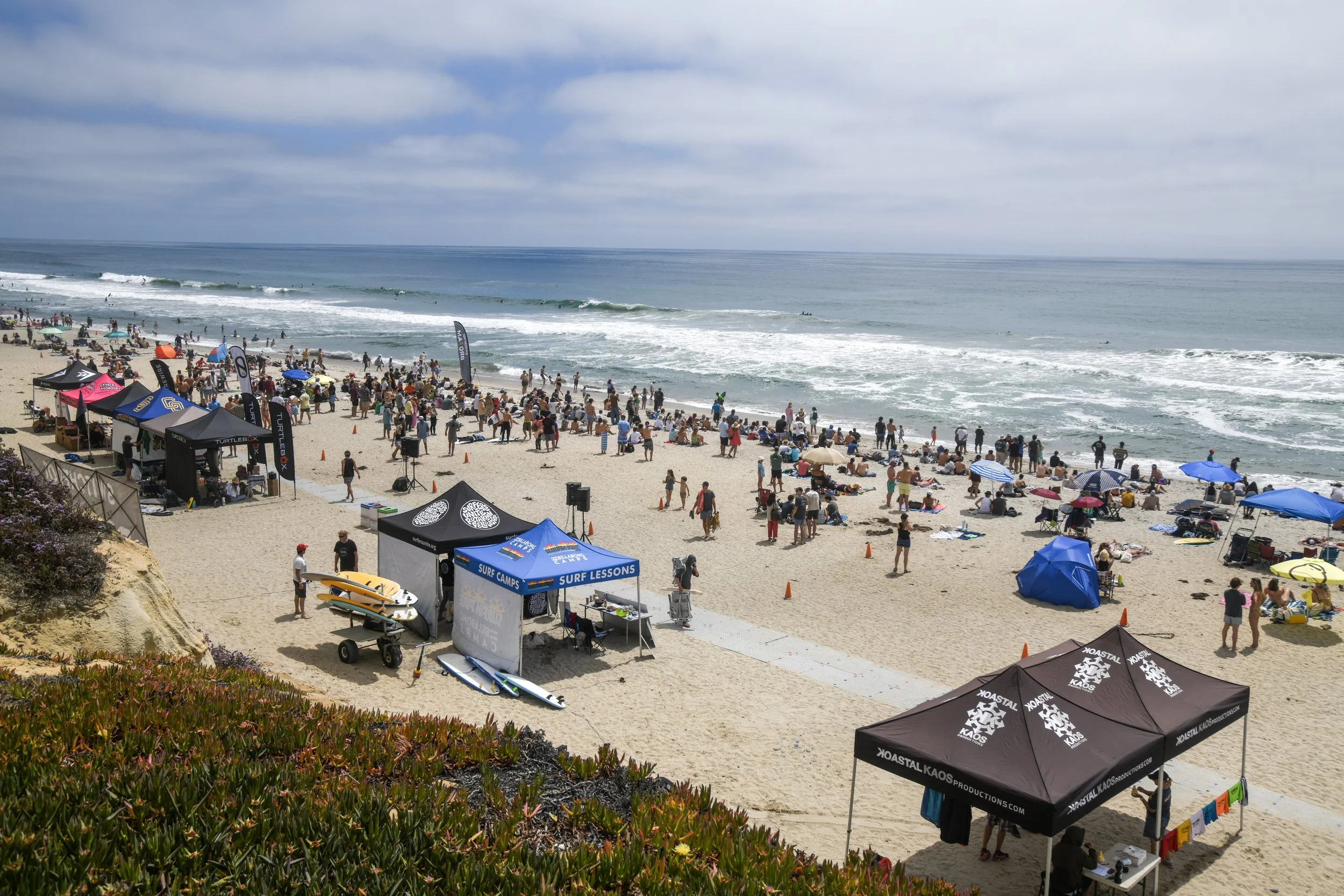 Crowded beach scene with surf lesson tents, beachgoers, and waves rolling in under a cloudy sky.