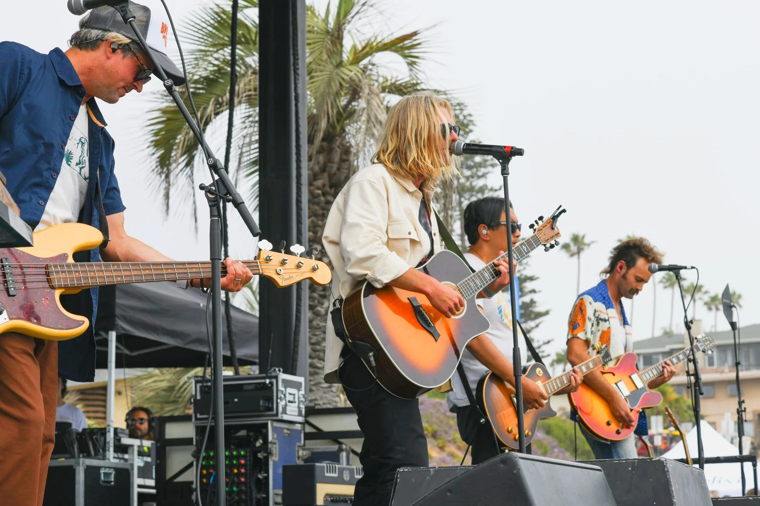 Outdoor live music performance featuring a singer playing acoustic guitar with bandmates in the background.
