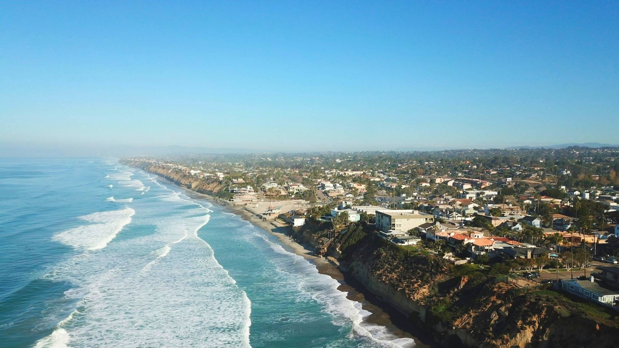 Aerial view of a coastal town with ocean waves crashing along cliffs and shoreline homes.