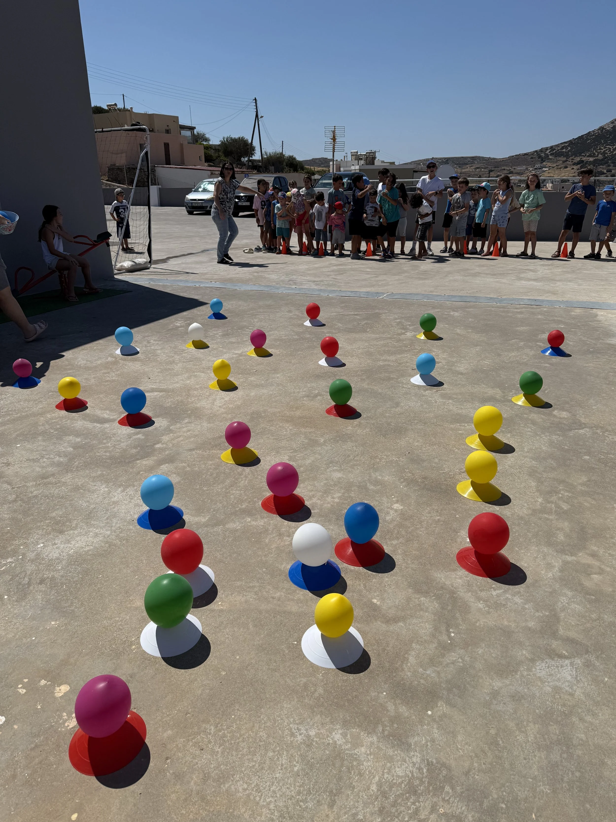 Children and adults in line on a sunny day for an outdoor activity, with colorful balls placed on the ground in the foreground, and houses and hills in the background.