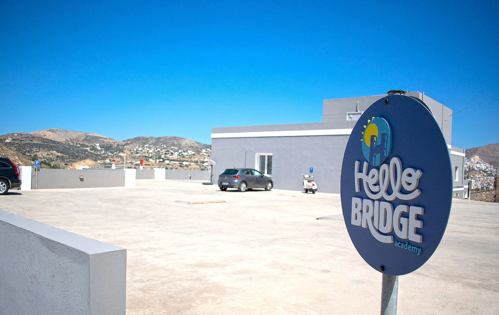 Exterior view of Hello Bridge Academy in Syros with the school sign, parking area, and surrounding island landscape.