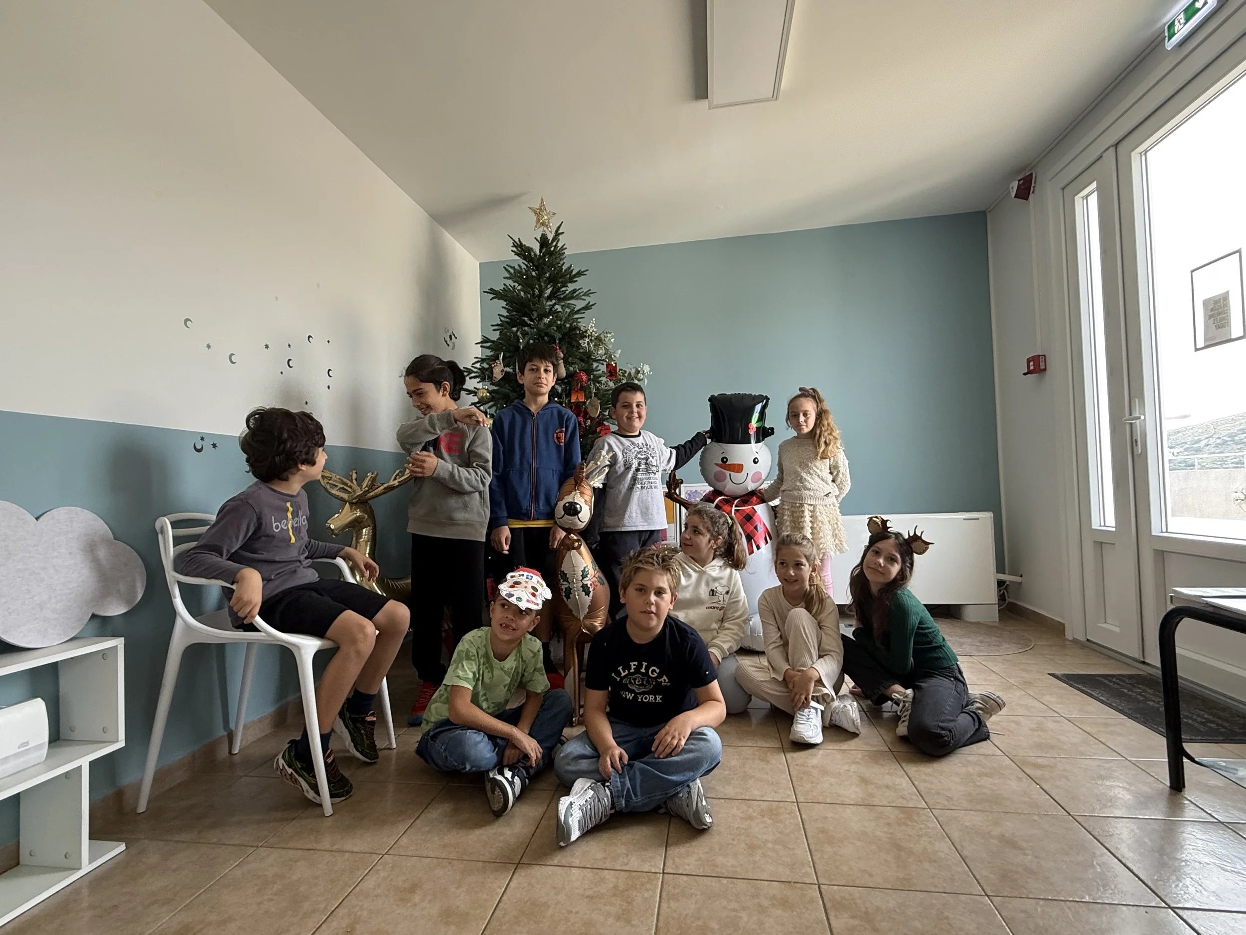 Group of children and a woman in front of a decorated Christmas tree, some children wearing reindeer antler headbands, in a room with light blue and white walls and a large glass door.
