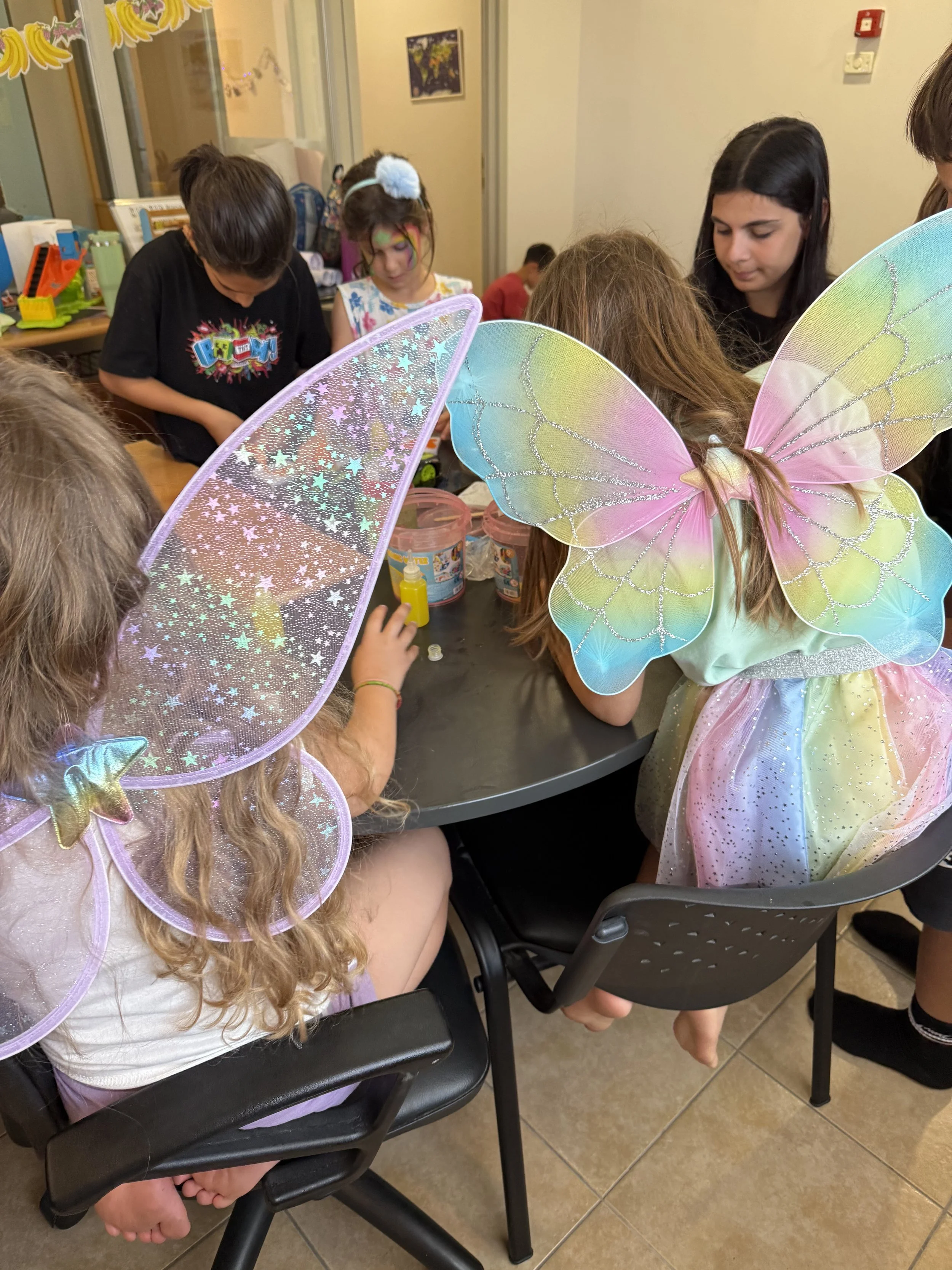 Children dressed as fairies with colorful wings gathered around a table, engaging in a craft activity in an indoor setting.