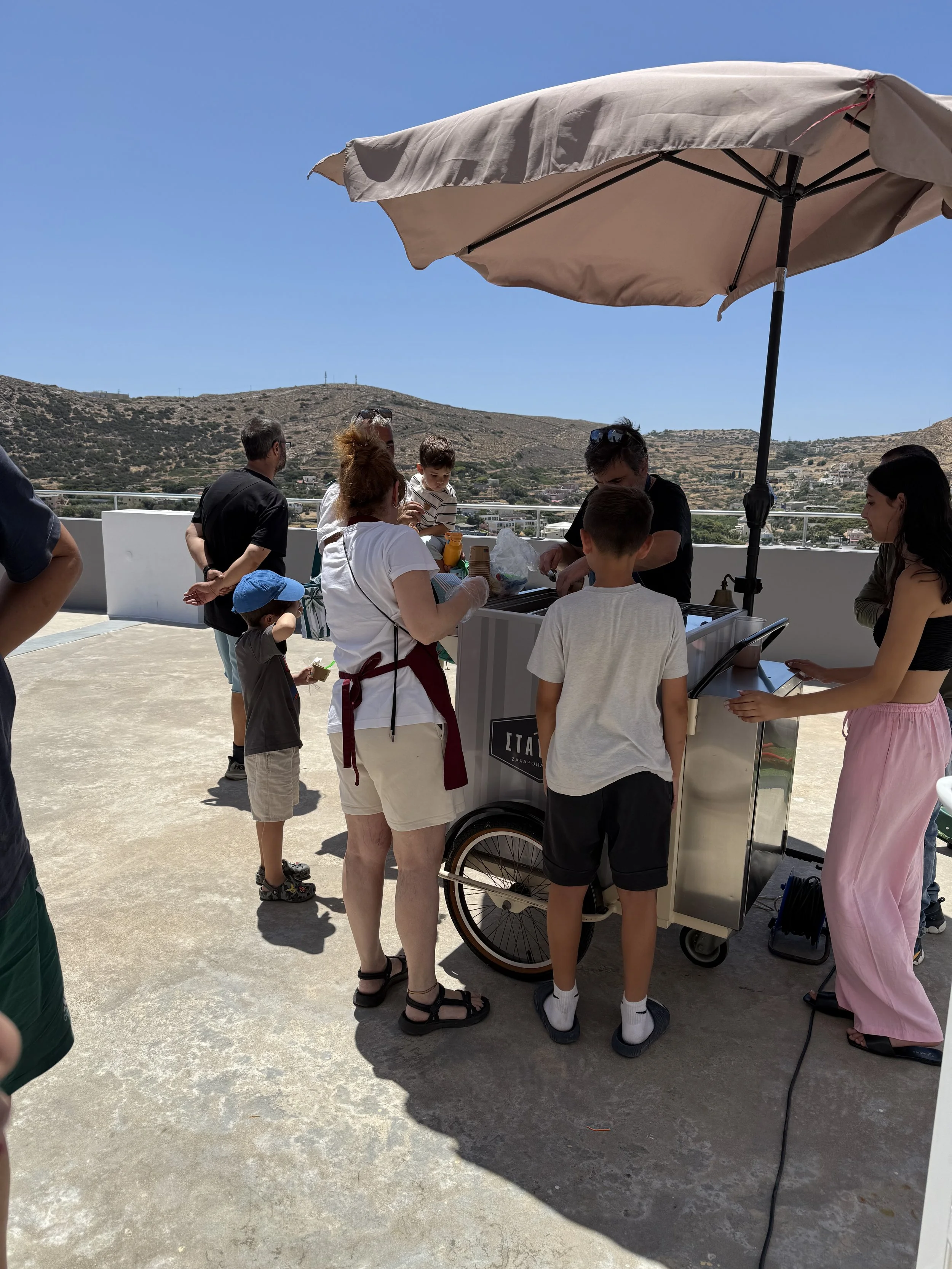 People gathering around a white ice cream cart on a rooftop terrace with hills in the background, under a large beige umbrella on a sunny day.