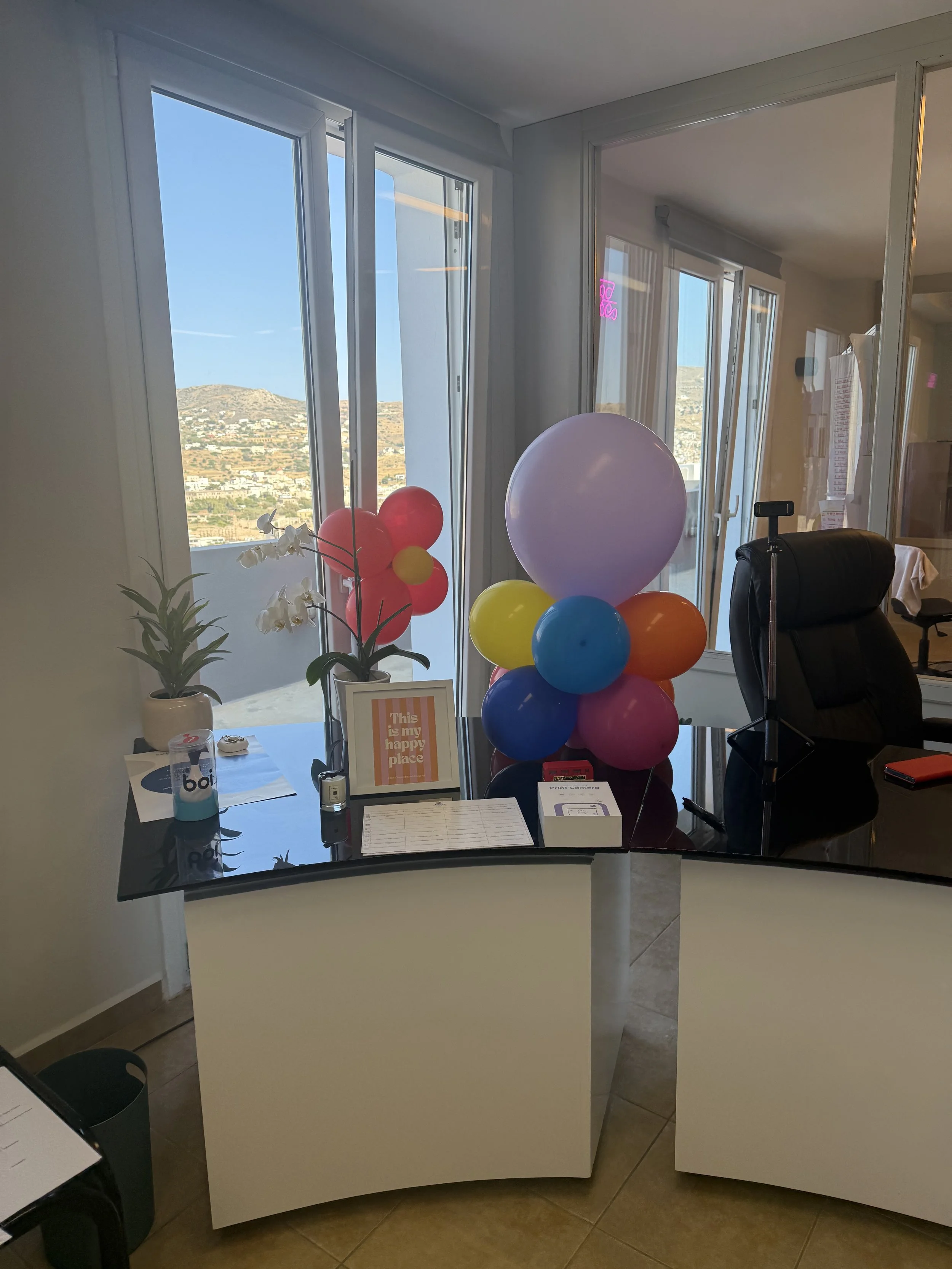 Office reception desk with balloons, a plant, and a sign that reads "This is my happy place." Behind the desk are windows showing a hillside landscape.