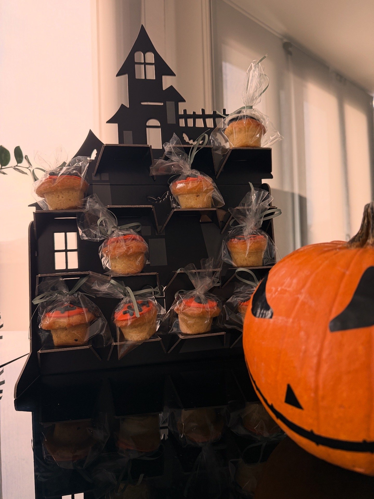 Black Halloween-themed display rack with small cupcakes wrapped in clear plastic bags tied with green ribbons, topped with orange and black icing, decorated for Halloween. A carved pumpkin with a black face is in the foreground.