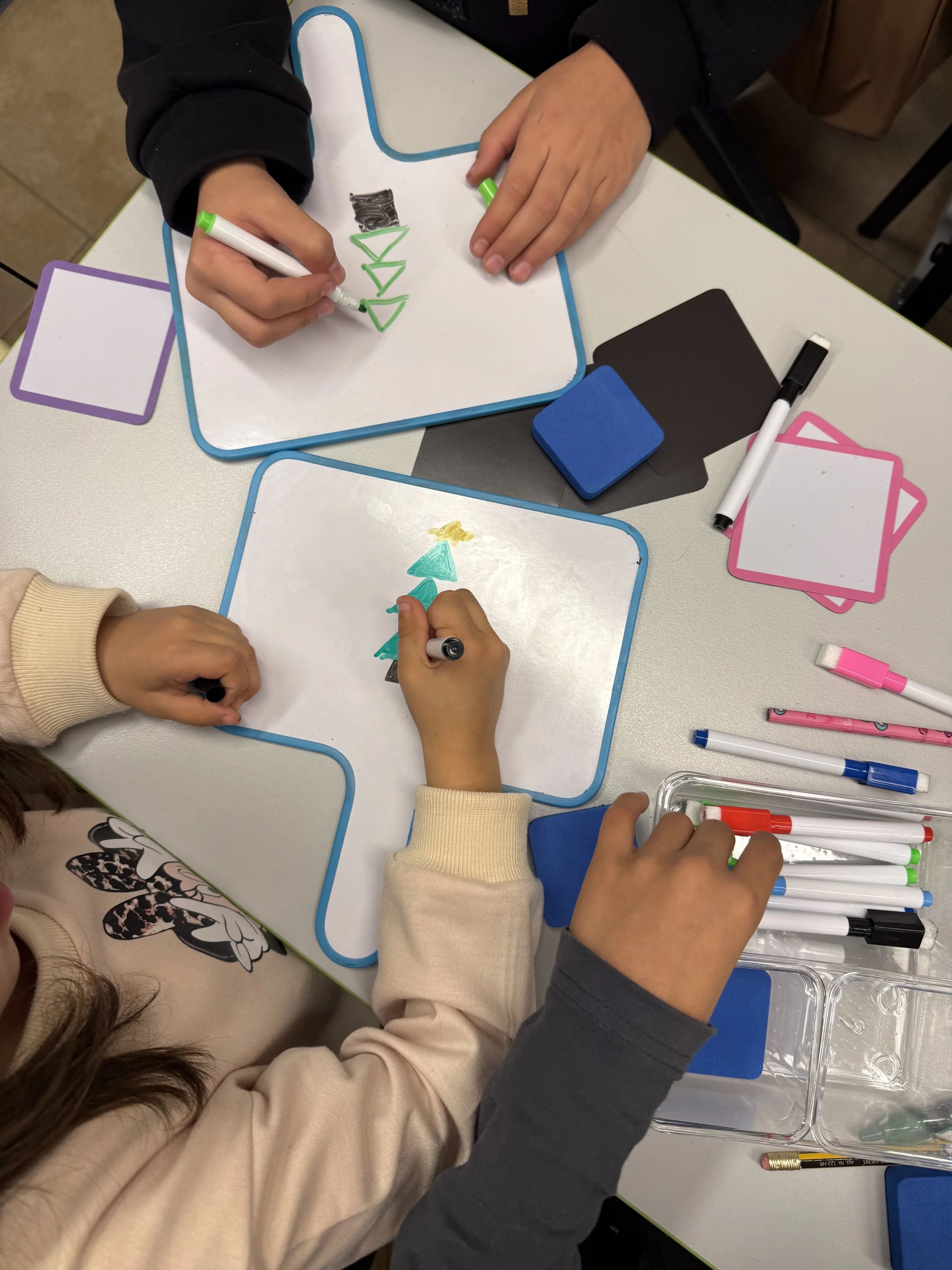 Children drawing Christmas trees and the word 'DAY' on whiteboards with markers.