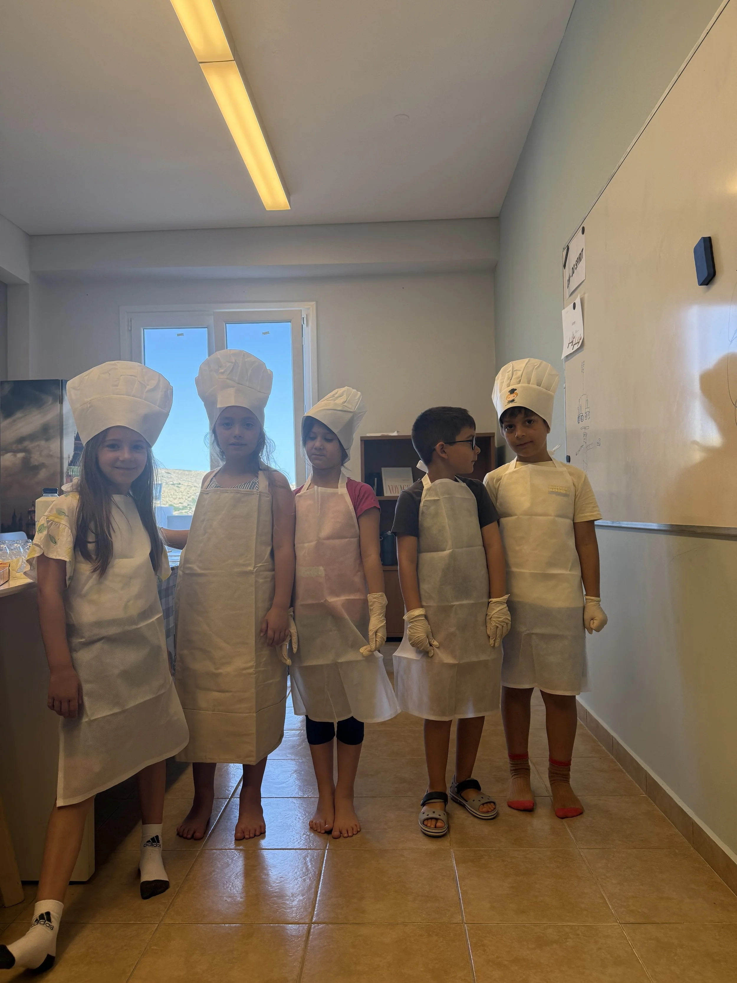 Children dressed as bakers standing in a classroom, wearing white aprons and chef hats, with some barefoot and others wearing socks or sandals.