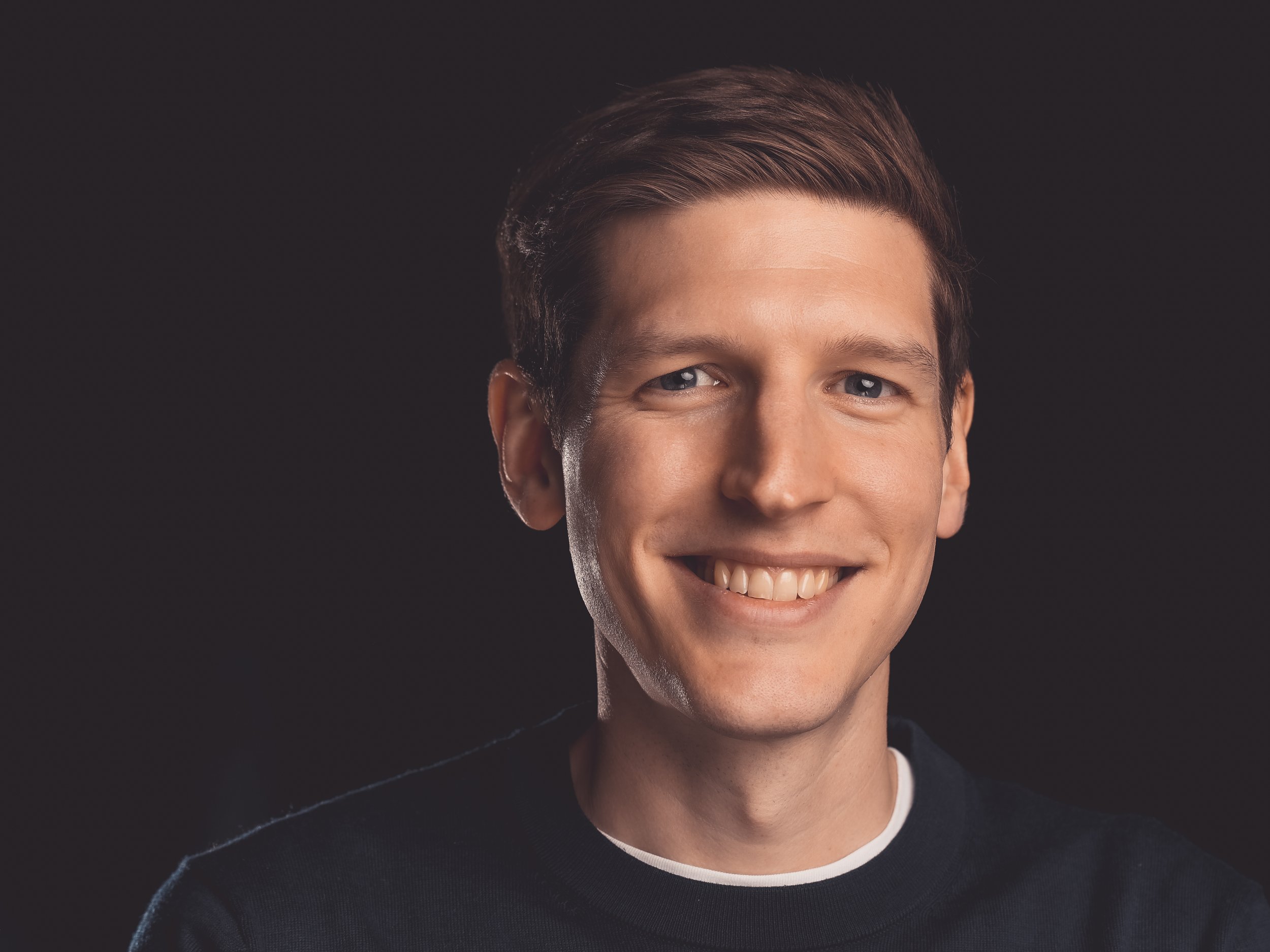 Close-up of a young man with short brown hair and blue eyes, smiling against a dark background.