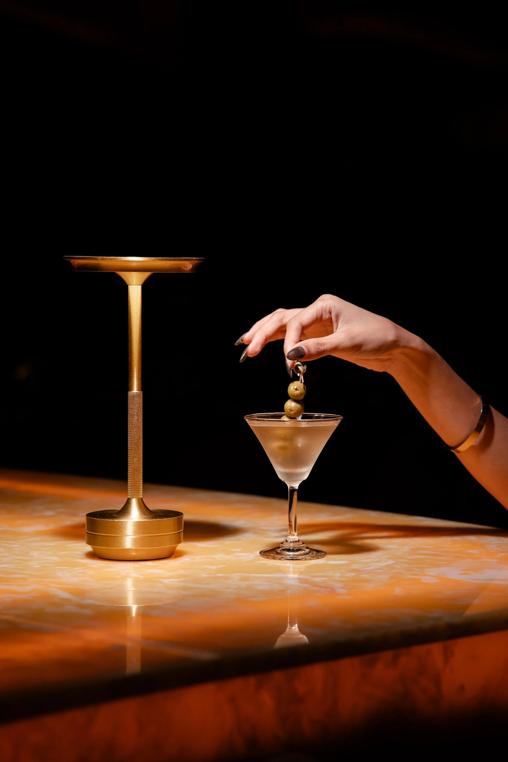 A hand is holding a skewer with three olives over a martini glass filled with a light-colored drink, on a marble surface, with a gold candlestick nearby, against a dark background.