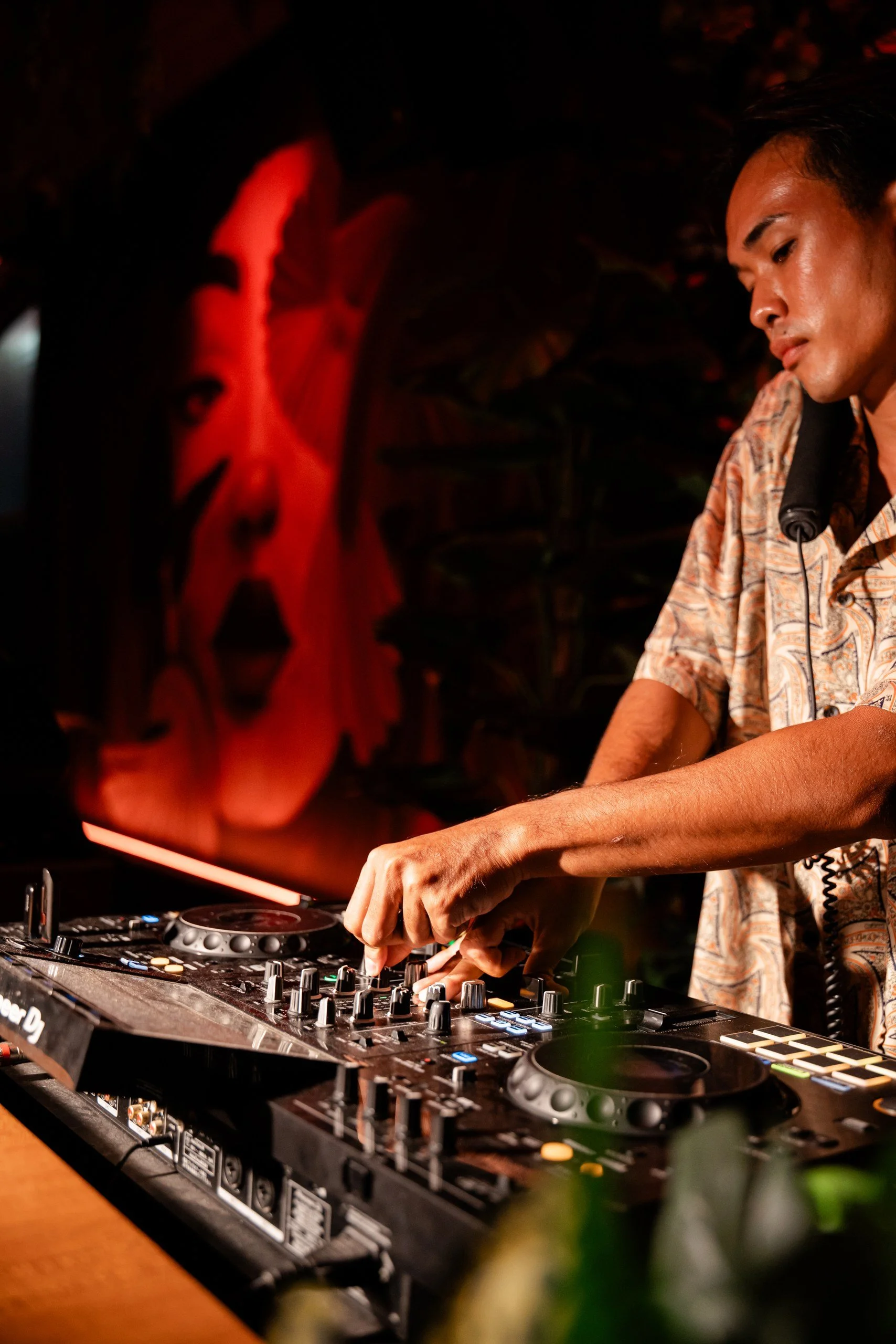 A person DJing at a party or club, wearing a patterned shirt, with turntables and a mixer, illuminated by red lighting and a large face mural in the background.