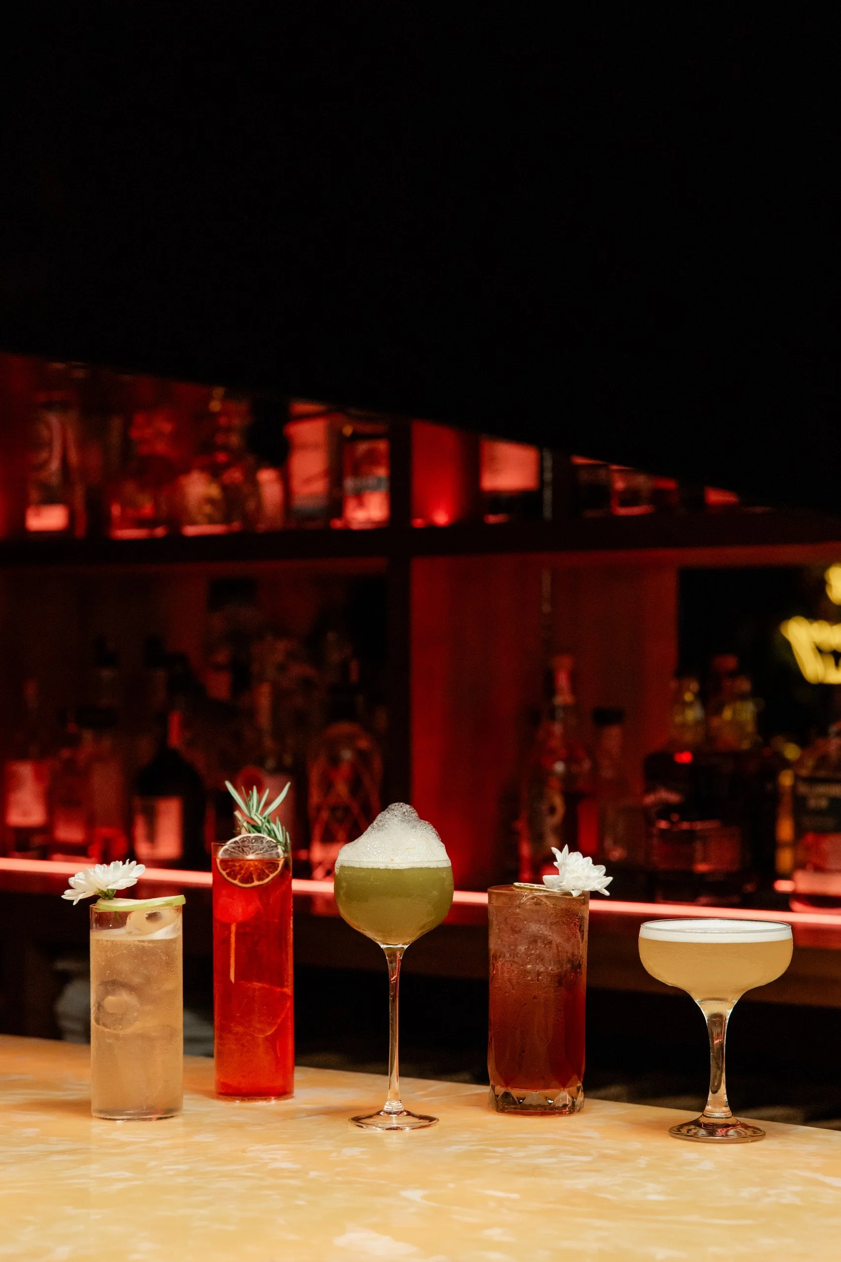 Five colorful cocktails on a bar counter with a dark bar background, each garnished with flowers, herbs, or slices of fruit.