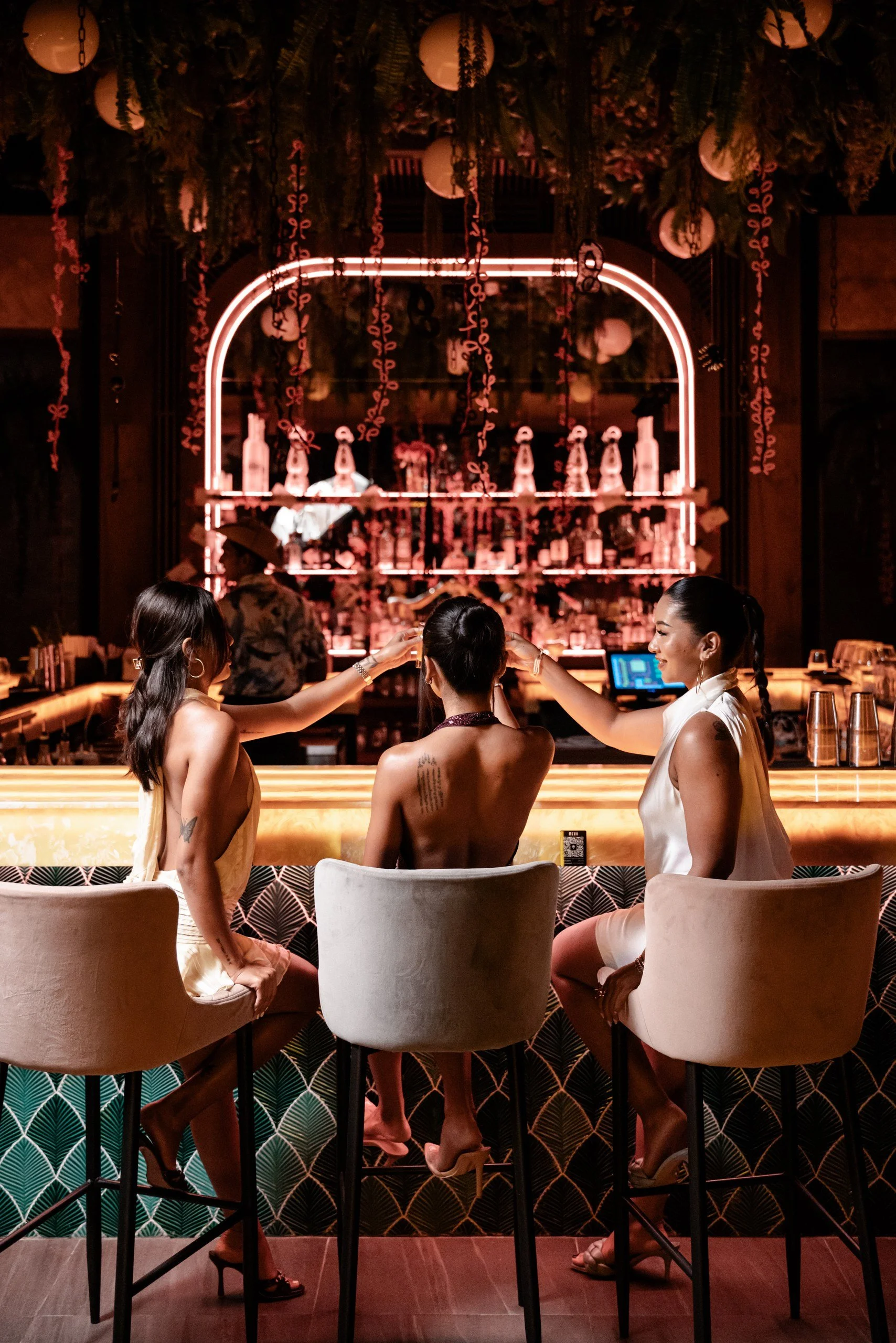 Three women sitting at a bar with a neon-lit back wall. One woman has long dark hair, another has her hair tied up, and the third has braided hair. They are wearing stylish dresses and high heels, raising glasses in a toast. The bar features decorative green patterned tiles and hanging floral arrangements.