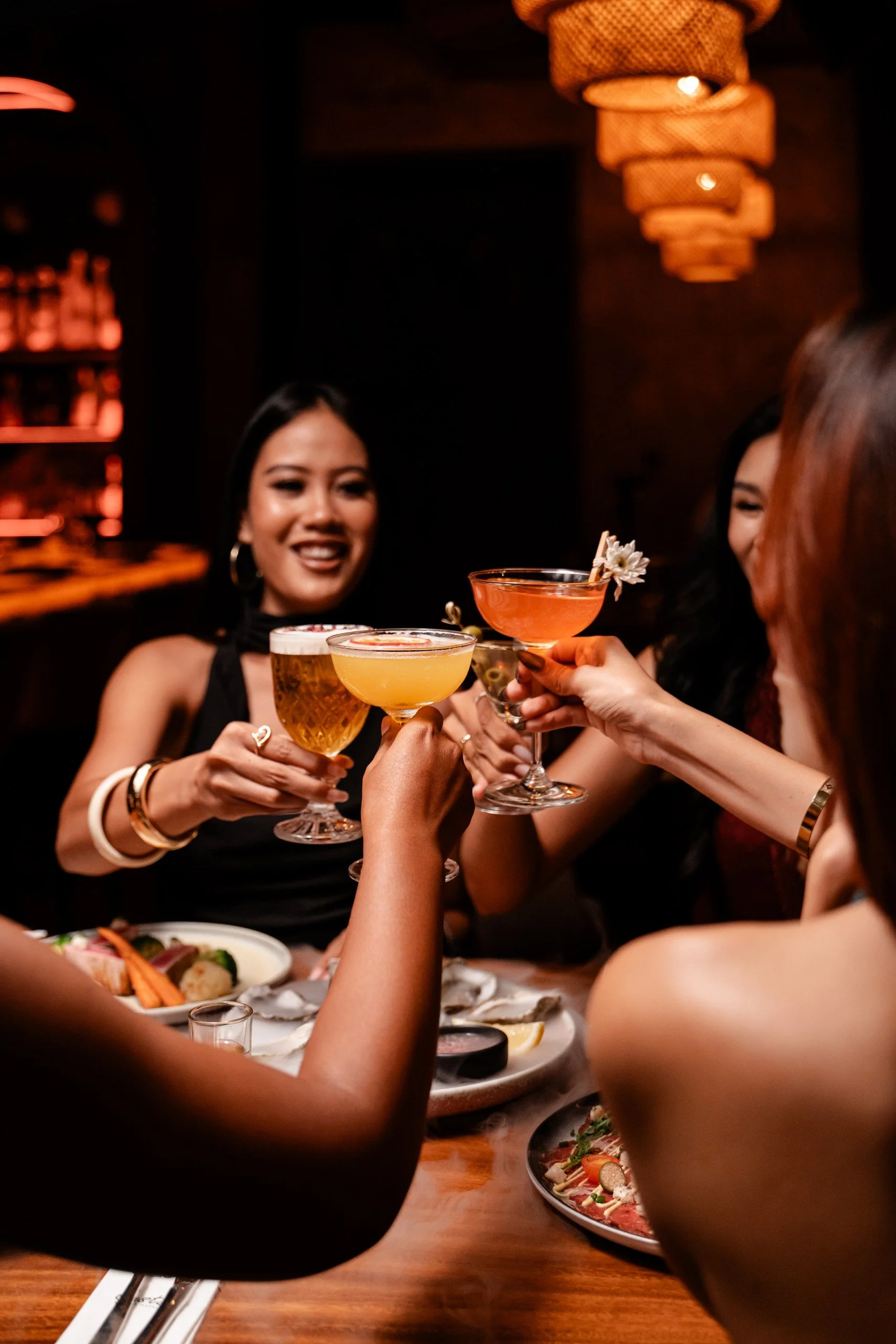 A group of women at a dinner table raising cocktails in a toast, with food and drinks visible on the table. The setting is warm and dimly lit.