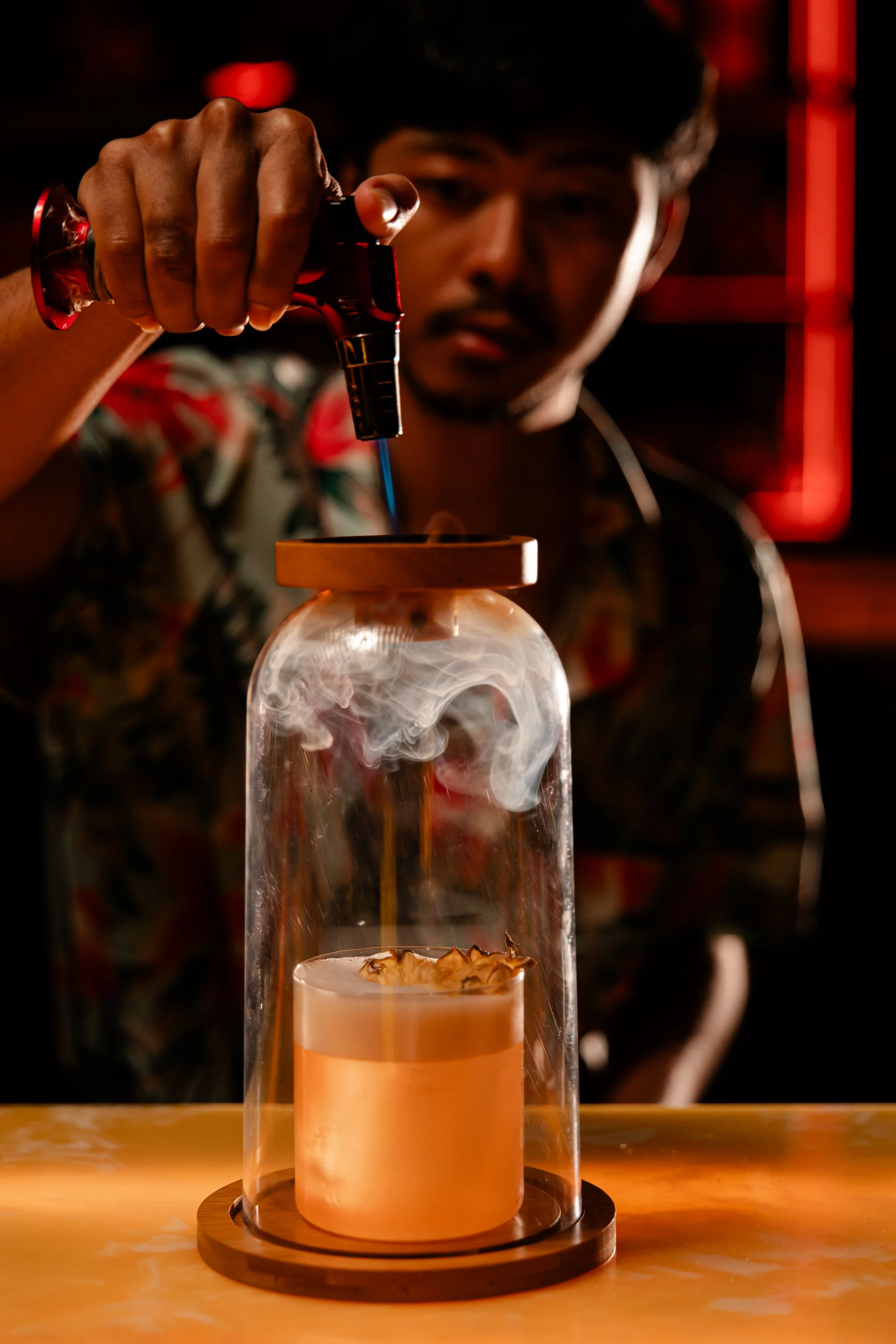 A person pouring liquid nitrogen into a glass container with smoke and vapor rising. The person is out of focus in the background, wearing a patterned shirt, in a dimly lit setting with red lighting.