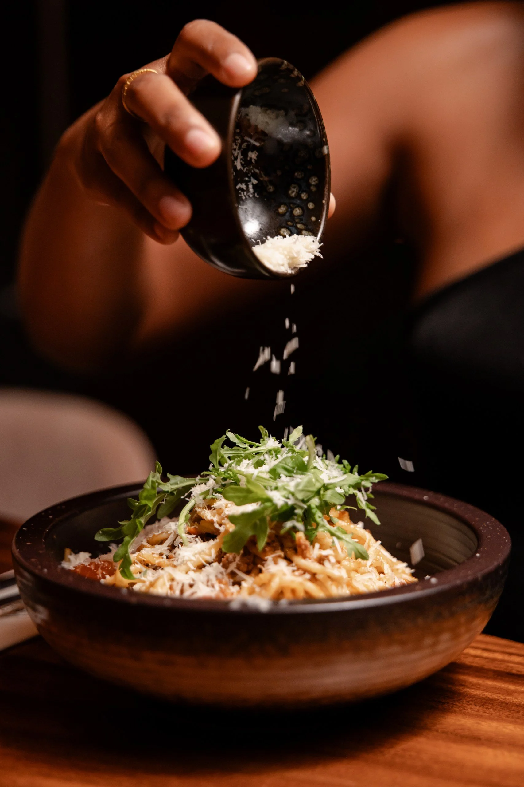 A person sprinkling cheese over a bowl of spaghetti with greens, on a wooden table.