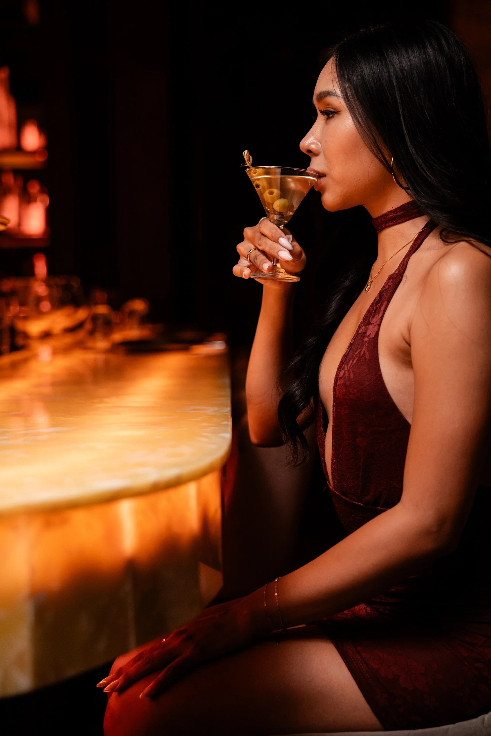 A woman with black hair and hoop earrings, wearing a burgundy lace dress and choker, sitting at a bar, holding a martini glass with olives, in a dimly lit setting.