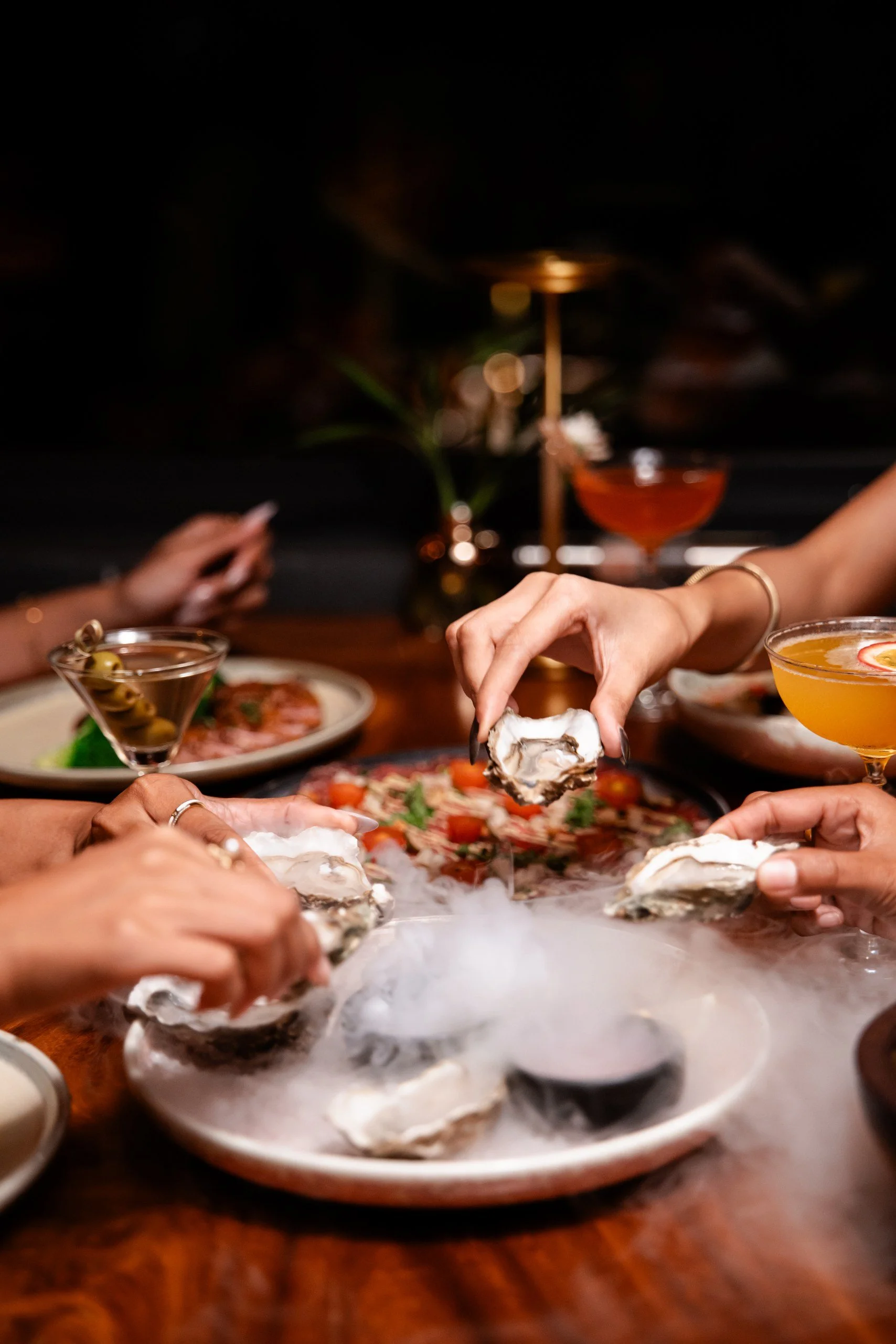 People gathered around a table eating oysters from a central platter with dry ice, with cocktails and appetizers visible on the table.