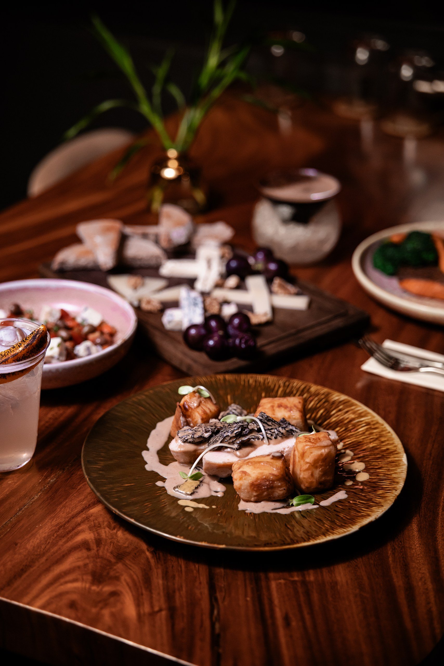 Close-up of a plated gourmet dish with fish, served on a decorative plate on a wooden table, with other dishes and a plant in the background.