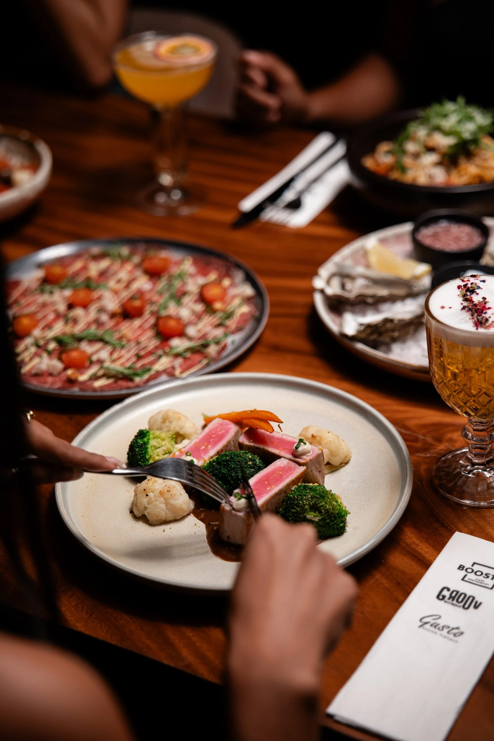 People sharing a meal at a restaurant, with various dishes and drinks on a wooden table, including a plate with broccoli, cauliflower, and pink meat, and a glass of beer with foam and flower petals