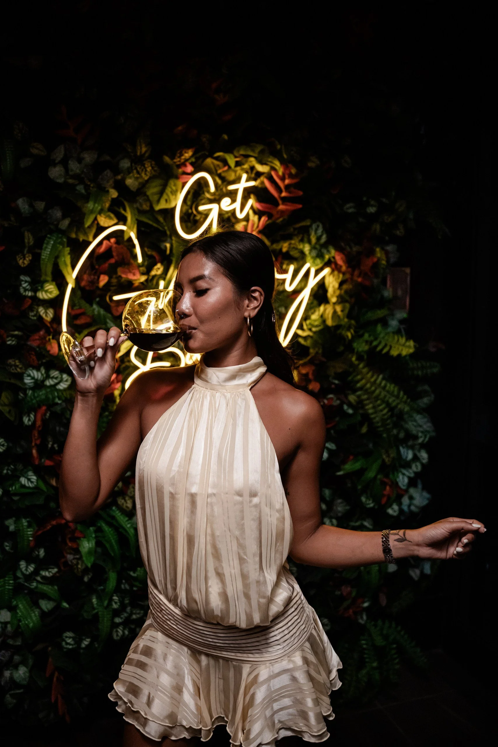 A woman in a cream-colored dress with vertical stripes, holding a glass of red wine, standing in front of a neon sign and a lush green floral backdrop.