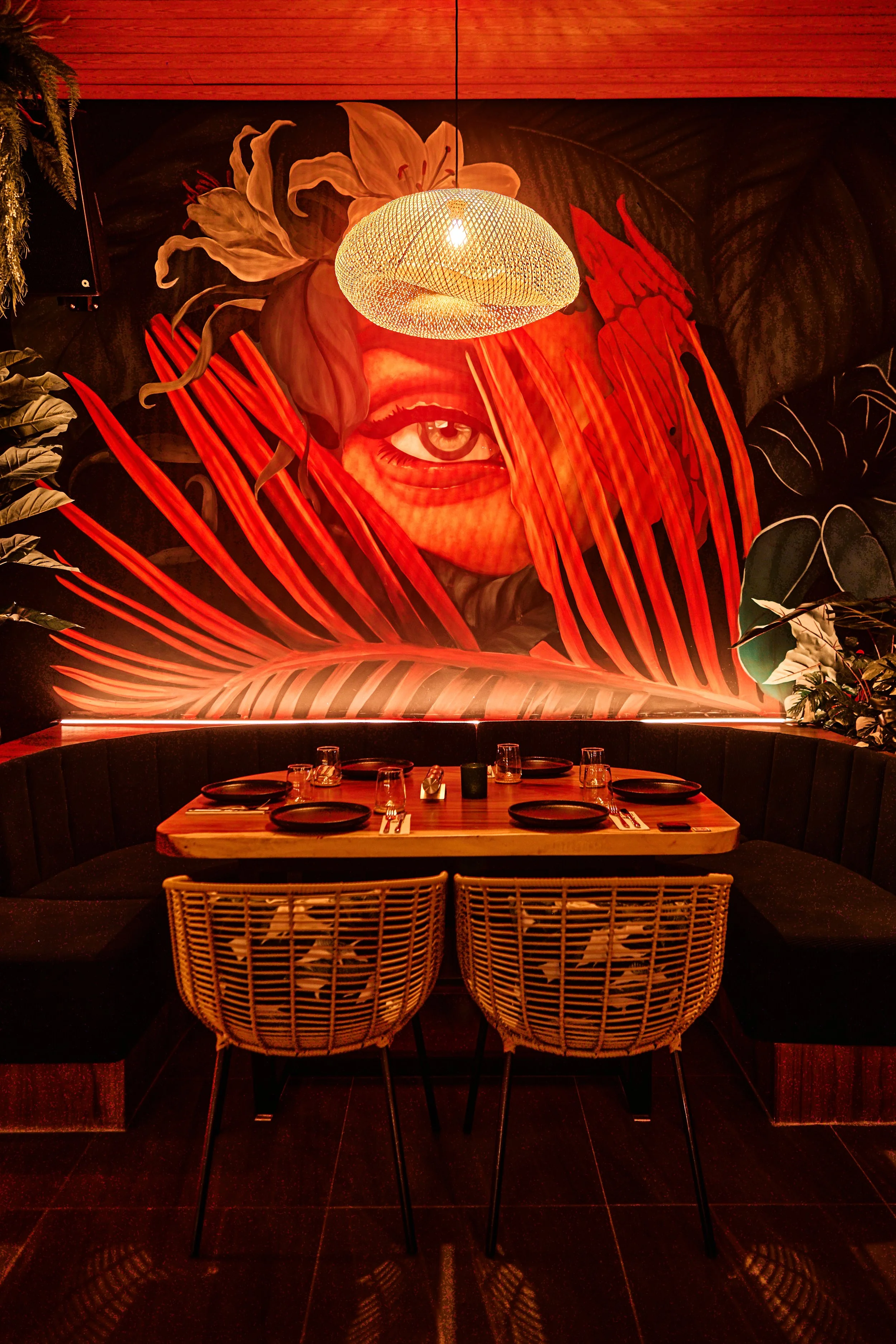 A restaurant dining area with a wooden table set for four, black plates, glasses, and utensils. Rattan chairs are positioned around the table. A large wall mural features a woman’s face partially obscured by red and orange tropical leaves, with a han
