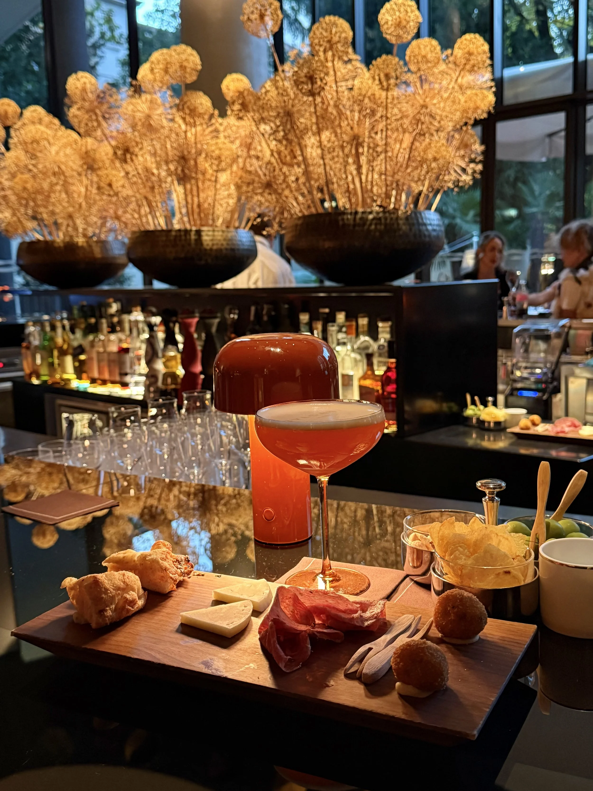 Orange Bulgari Cocktail beside a red lamp and an aperitivo board at the hotel bar, with large floral arrangements behind.