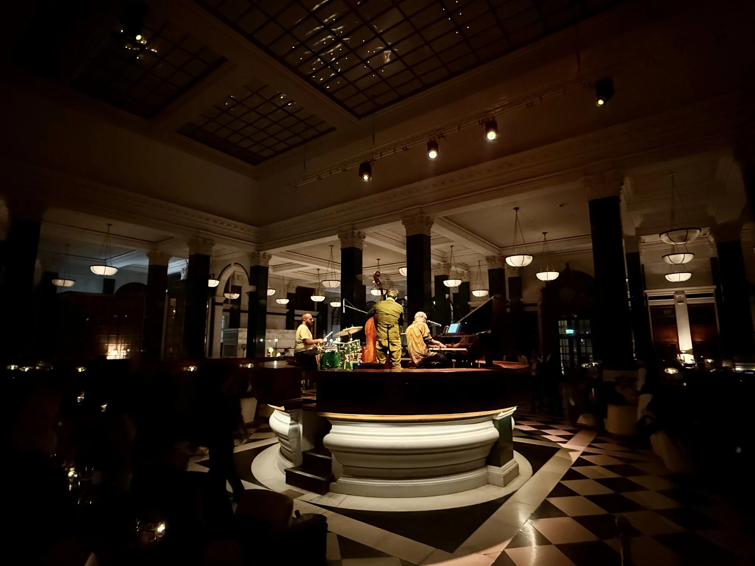 Three musicians playing drums, double bass and grand piano on a raised circular platform in The Ned’s dimly lit banking hall, surrounded by people beneath tall columns and pendant lights.