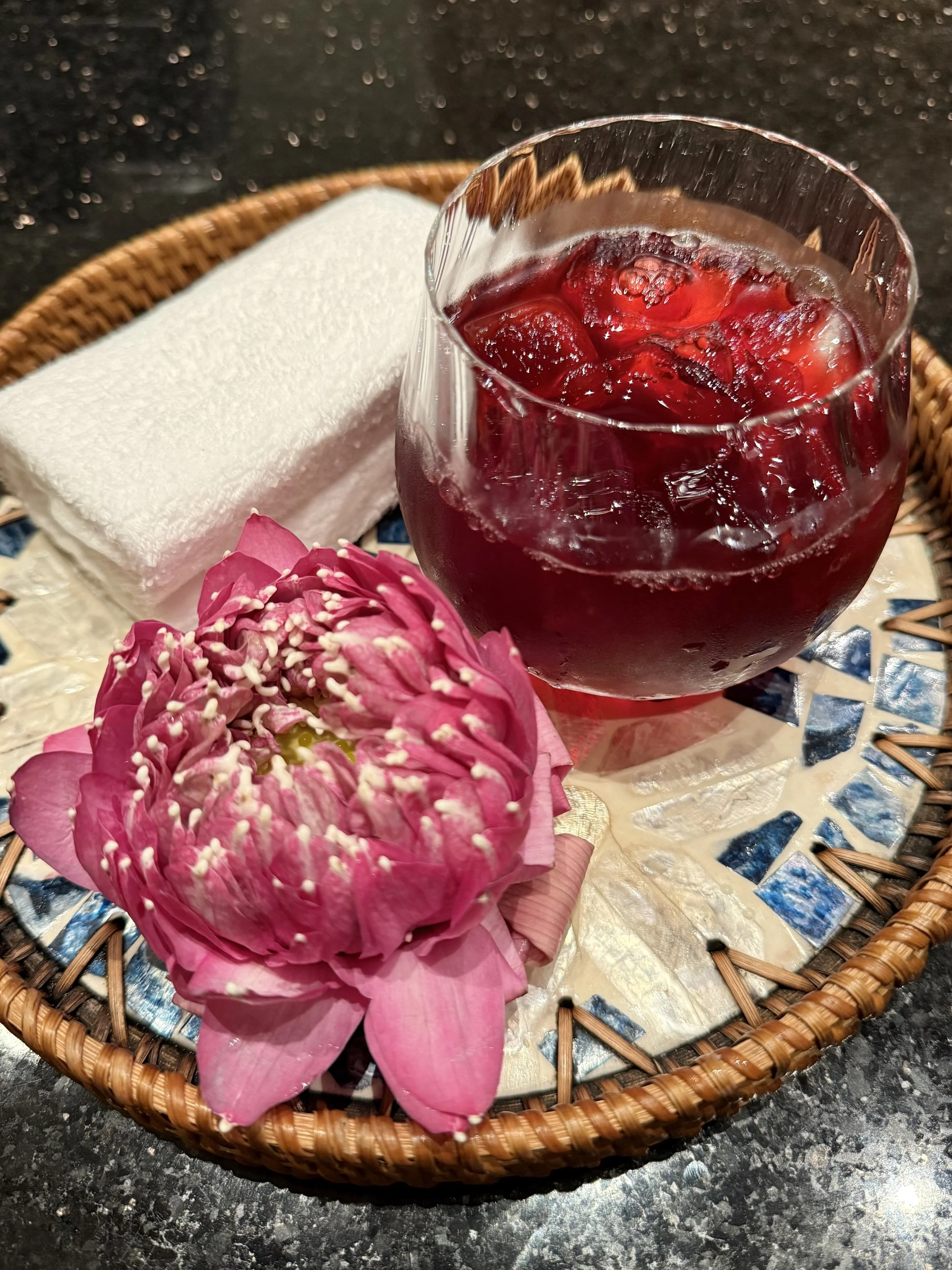A red iced welcome drink, white towel and folded pink lotus sit on a woven tray at The Sukhothai Bangkok.