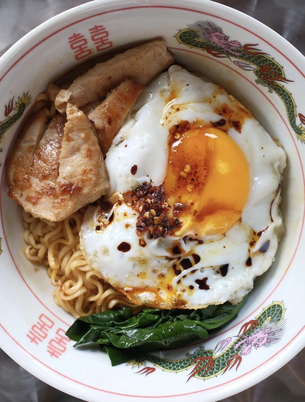 Close-up of noodles topped with sliced pork chop, a fried egg, chilli and leafy greens in a bowl decorated with dragons.