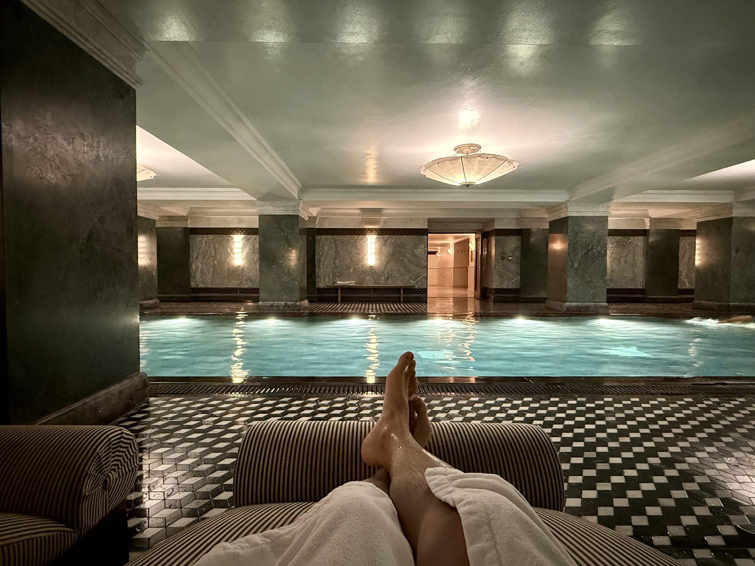 View from a striped lounger beside The Ned’s indoor pool, with towel-covered legs in the foreground, illuminated water, green columns, warm wall lights and geometric black-and-white flooring.