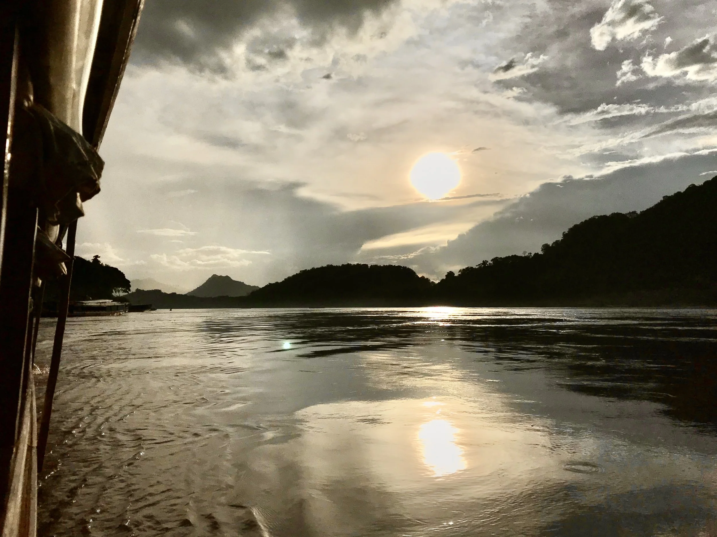 photograph taken from the boat at sunset: the sun sits above the dark mountain ridge, with a bright reflection across the wide Mekong and dramatic grey and gold clouds overhead.