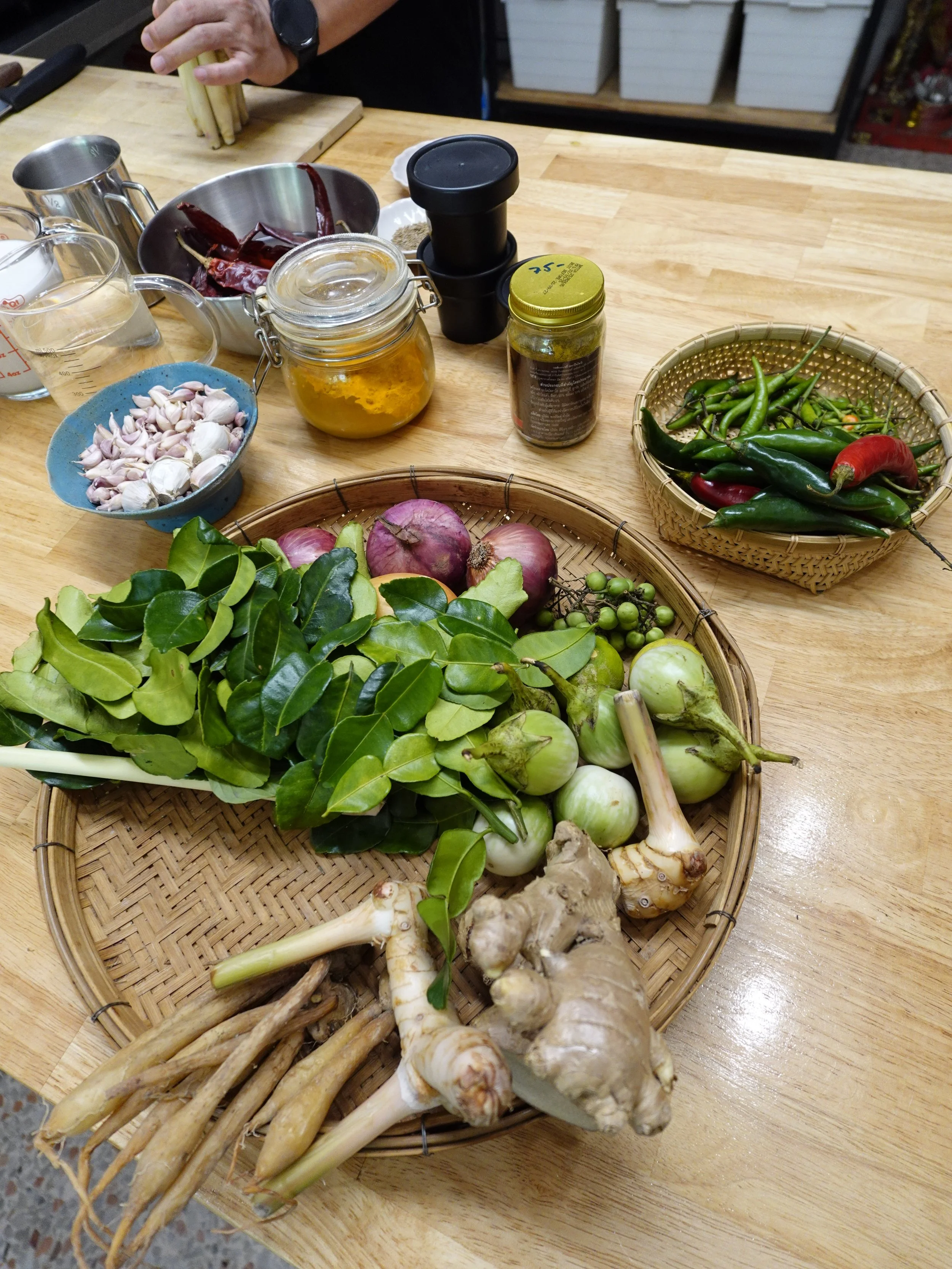 Woven trays of green aubergines, citrus leaves, roots and chillies beside garlic, dried red chillies and jars on a wooden counter.