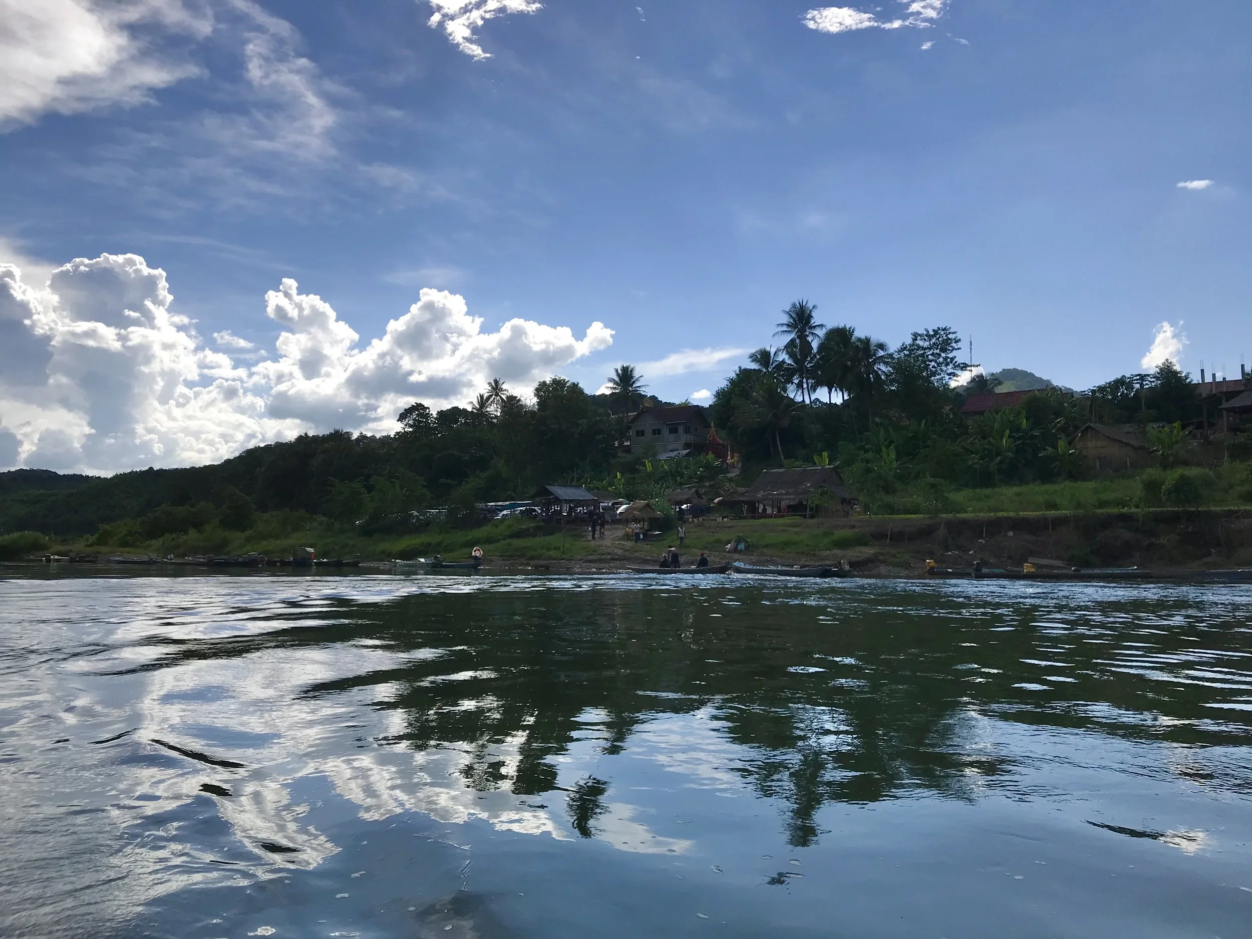 rippling dark water fills the foreground, a low green bank and a few small buildings sit across the river, and bright white clouds break across the blue sky.