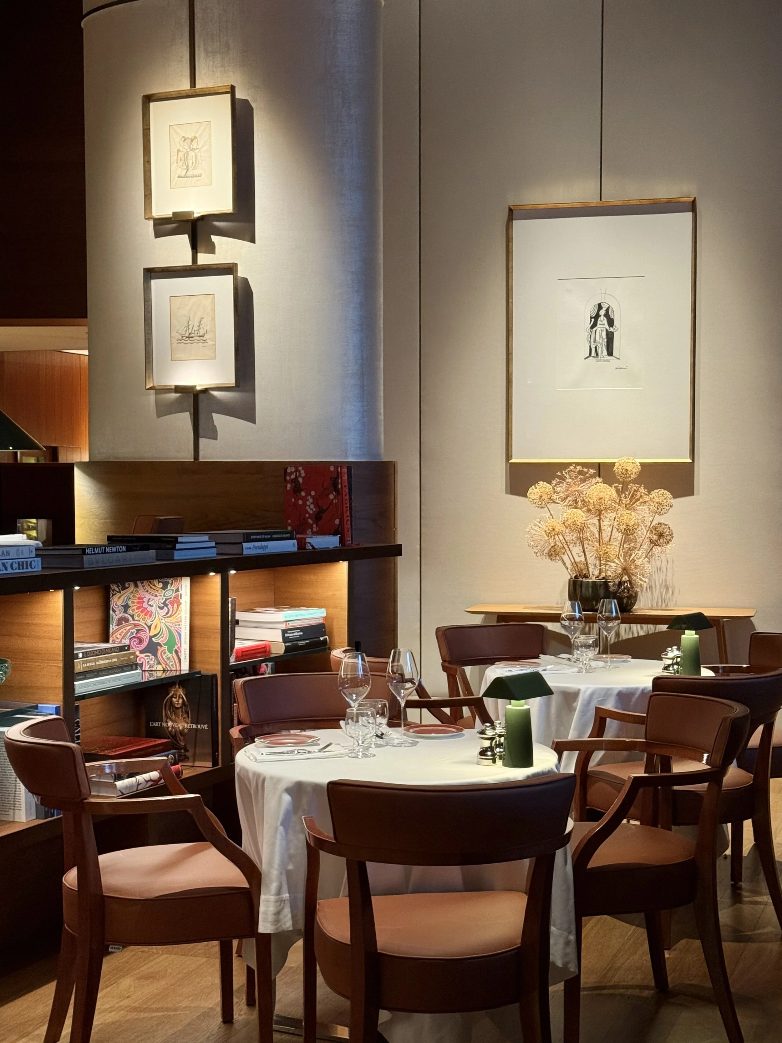 Small round tables with white cloths, green lamps and wooden armchairs beside bookshelves and framed drawings at Da Niko.