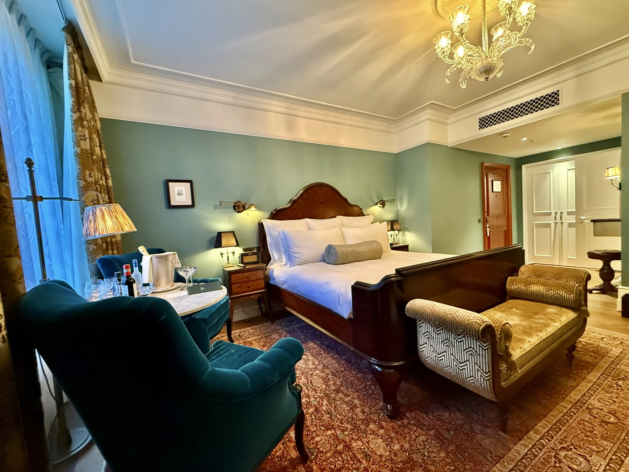 Medium Room at The Ned with green walls, a dark wooden bed, white bedding, teal armchairs, patterned curtains and rug, a gold-toned upholstered bench and a glass chandelier.