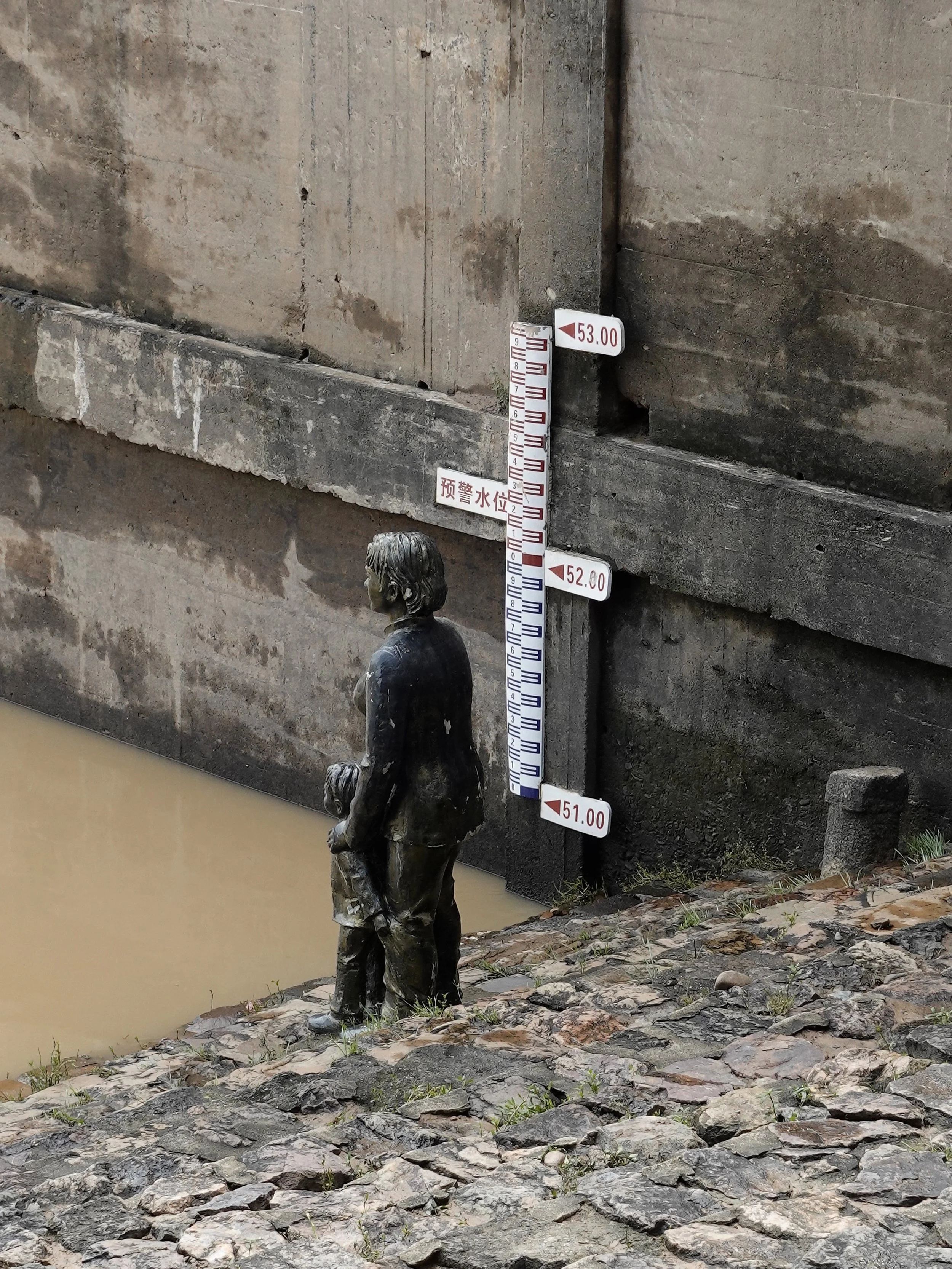 A sculpture of a woman with a child beside the stone steps at Huochuan Pier, with muddy river water below and a water-level gauge on the wall behind them.