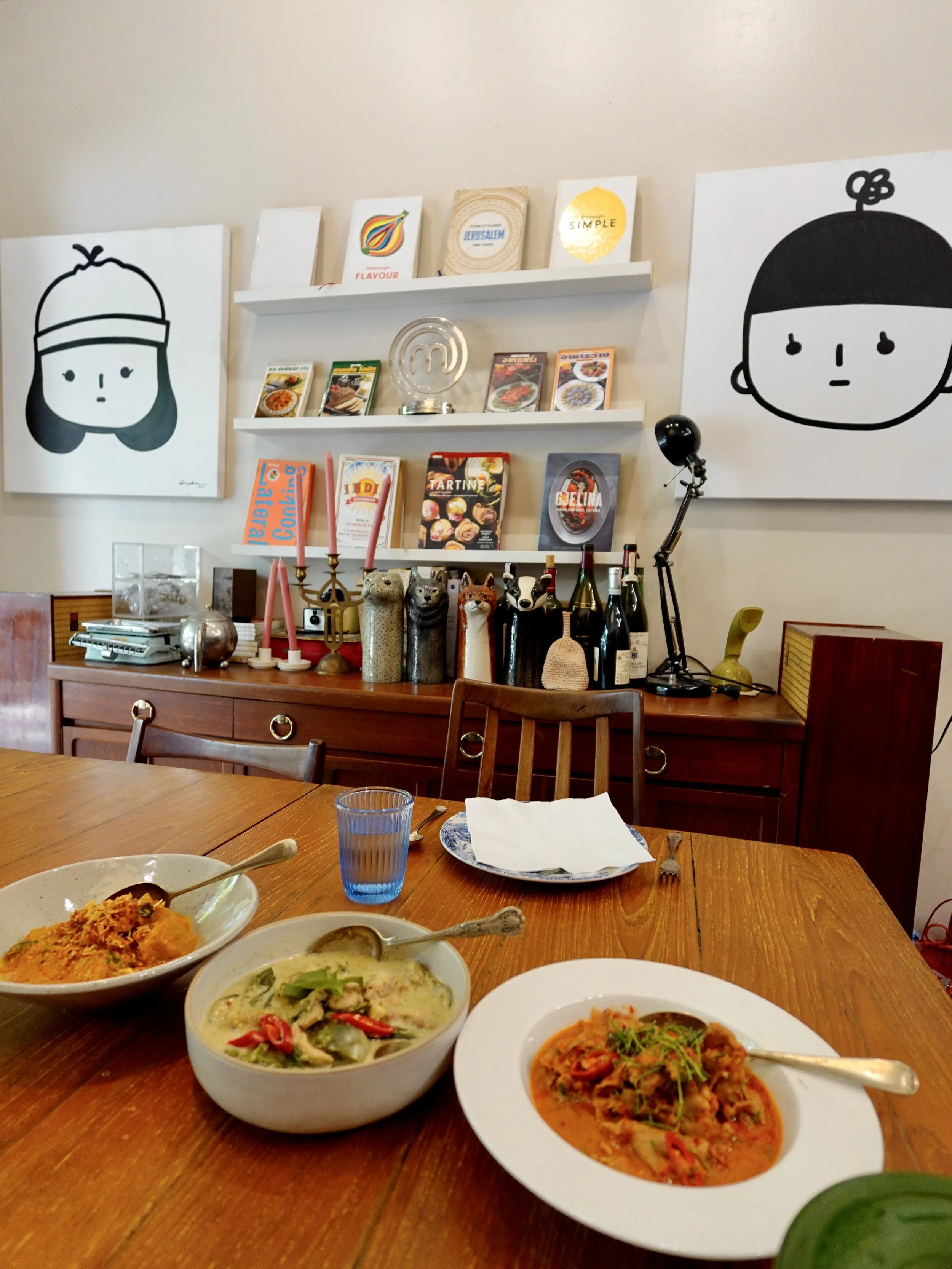 Three bowls of curry on a wooden dining table, with cookbooks, a MasterChef trophy, framed artwork and collected objects behind.
