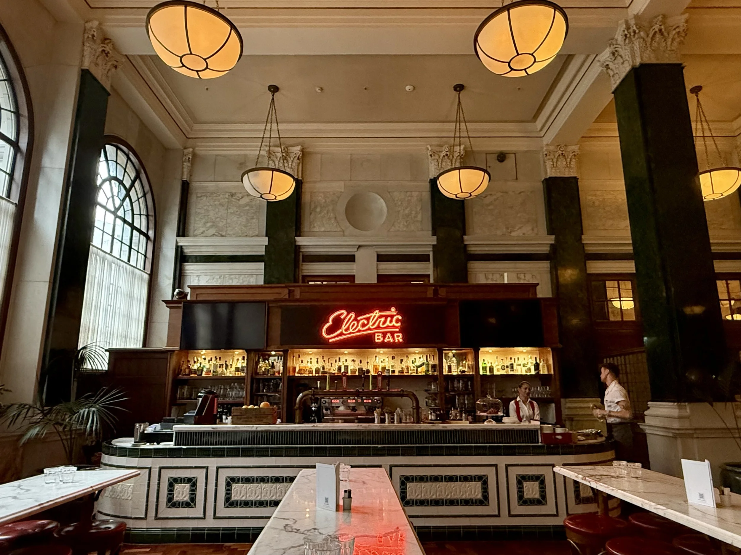 Electric Bar at The Ned, with a red neon sign, marble-topped tables, red stools, tall arched windows and warm pendant lights between green columns.
