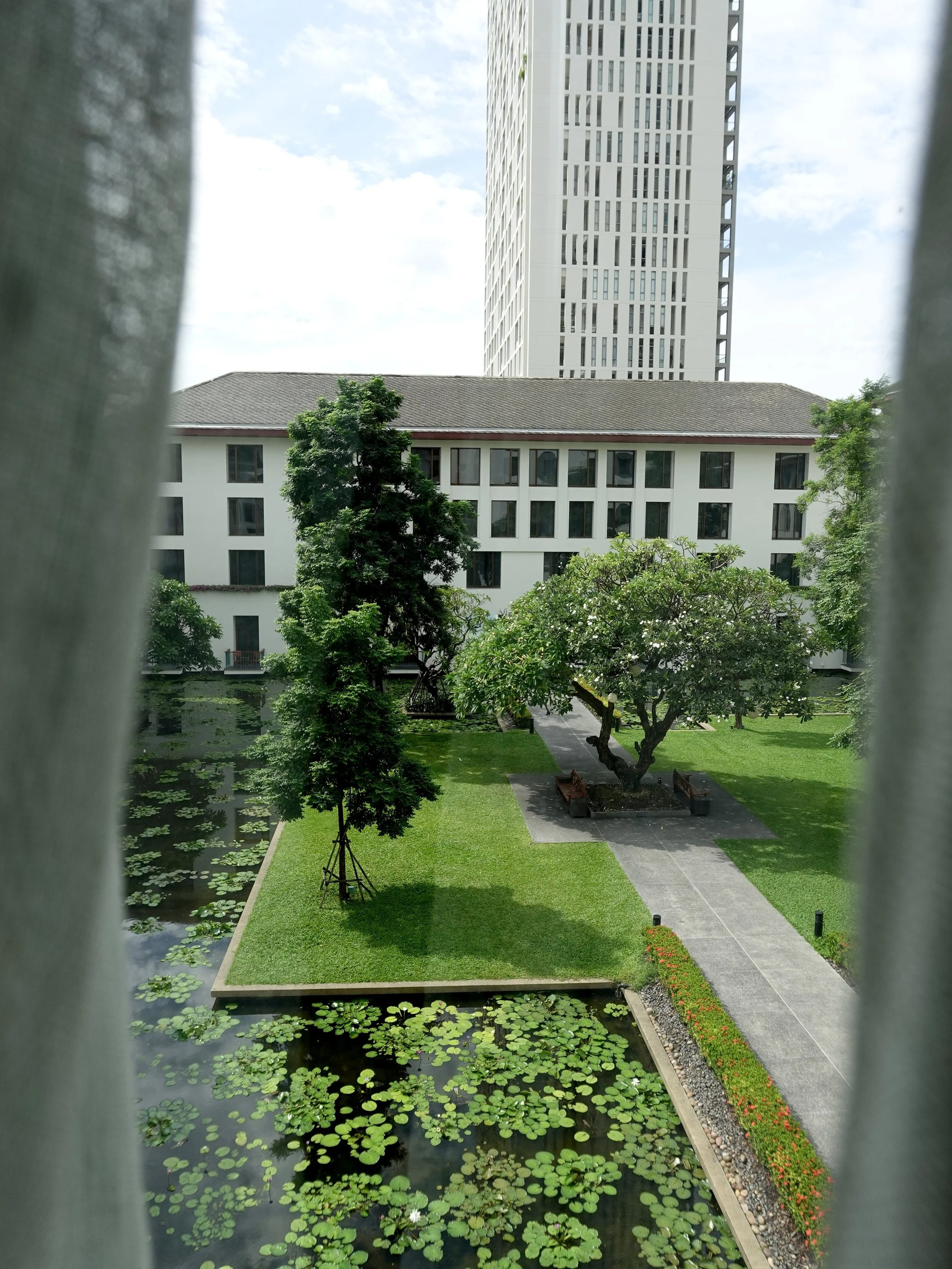 Curtains frame a view of courtyard trees, lawn and lily ponds, with a tall building beyond The Sukhothai Bangkok.