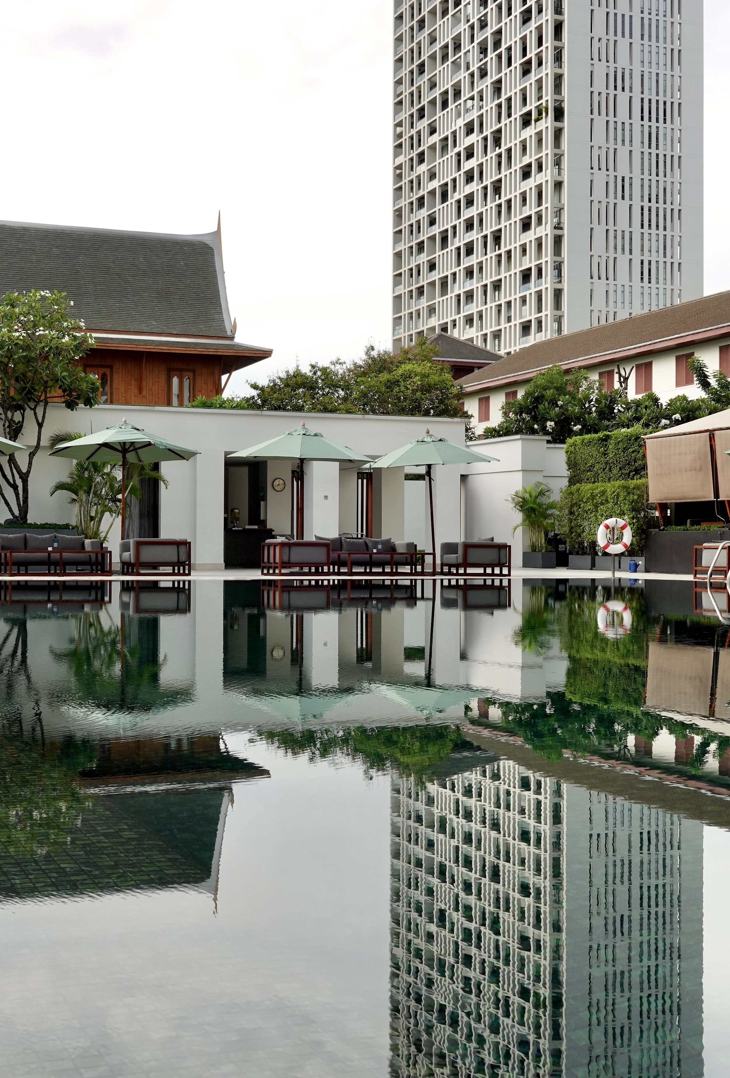 An empty swimming pool reflects green parasols, a Thai-style roof and a tall building at The Sukhothai Bangkok.