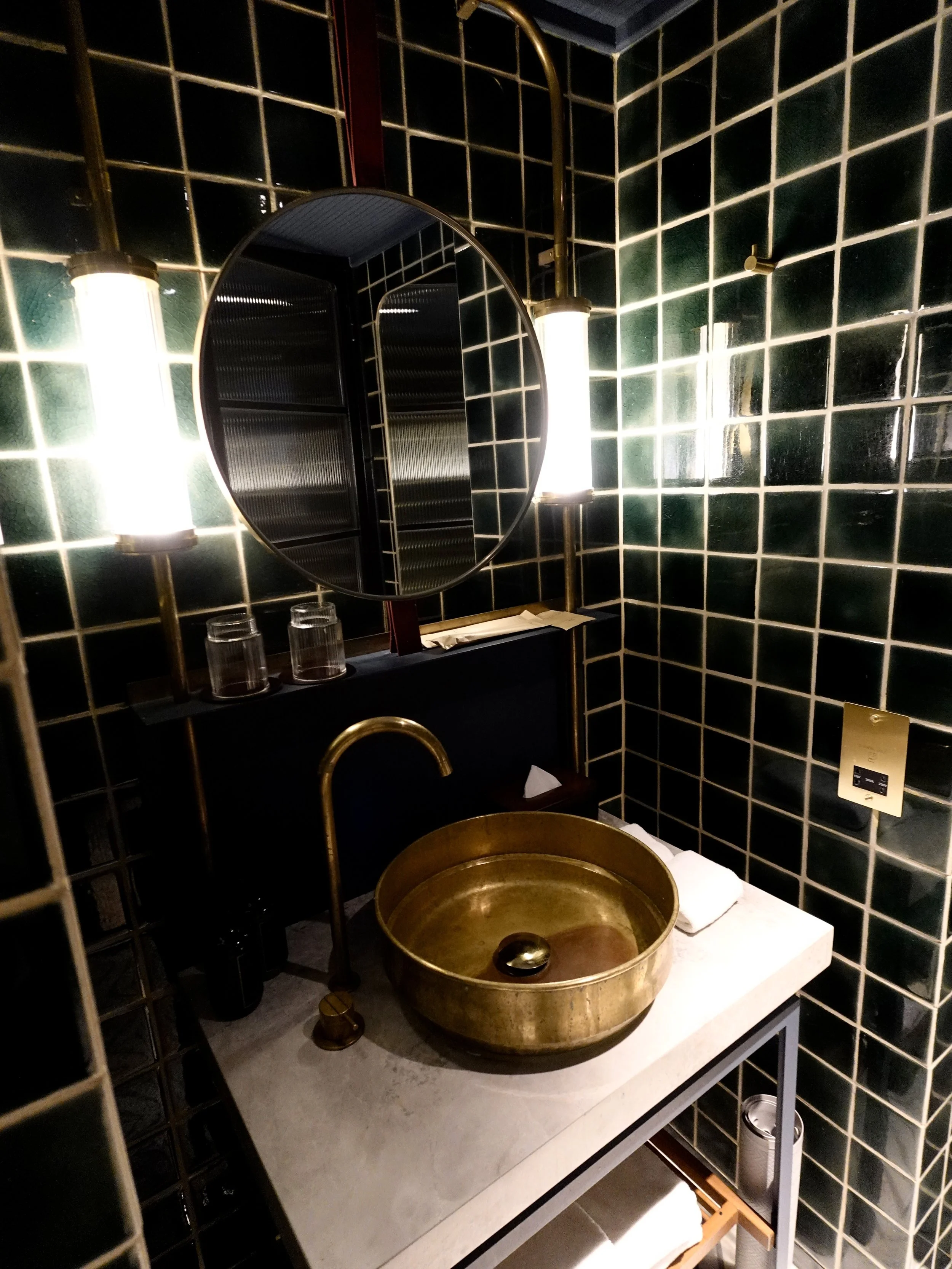 Bathroom at The Fleming with dark green ceramic tiles, round mirror, brass basin