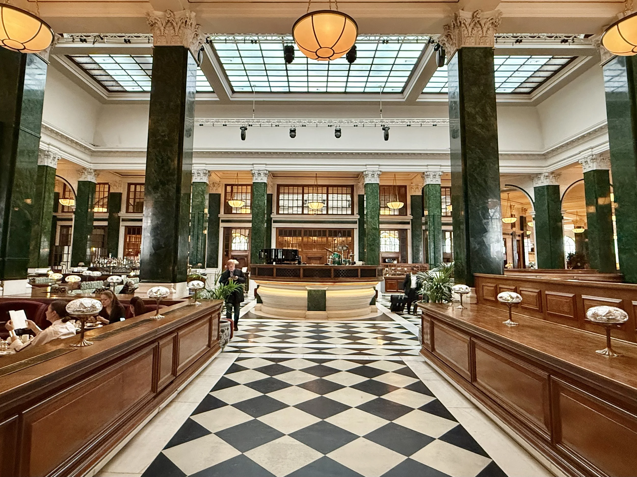 Symmetrical view of The Ned banking hall, with tall green columns, bowl-shaped pendant lights, long counters and a black-and-white diamond floor leading towards a clock above the doorway.