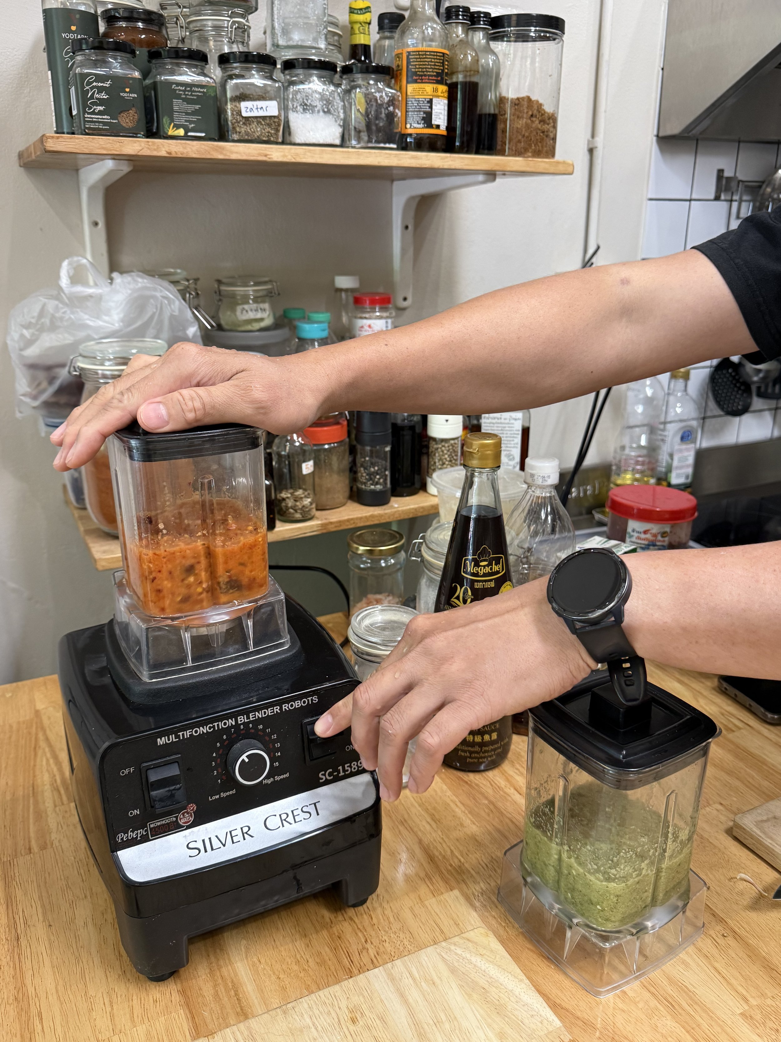 Angkrit’s hands operating a blender containing an orange-red curry mixture, with a separate jug of green mixture beside it.