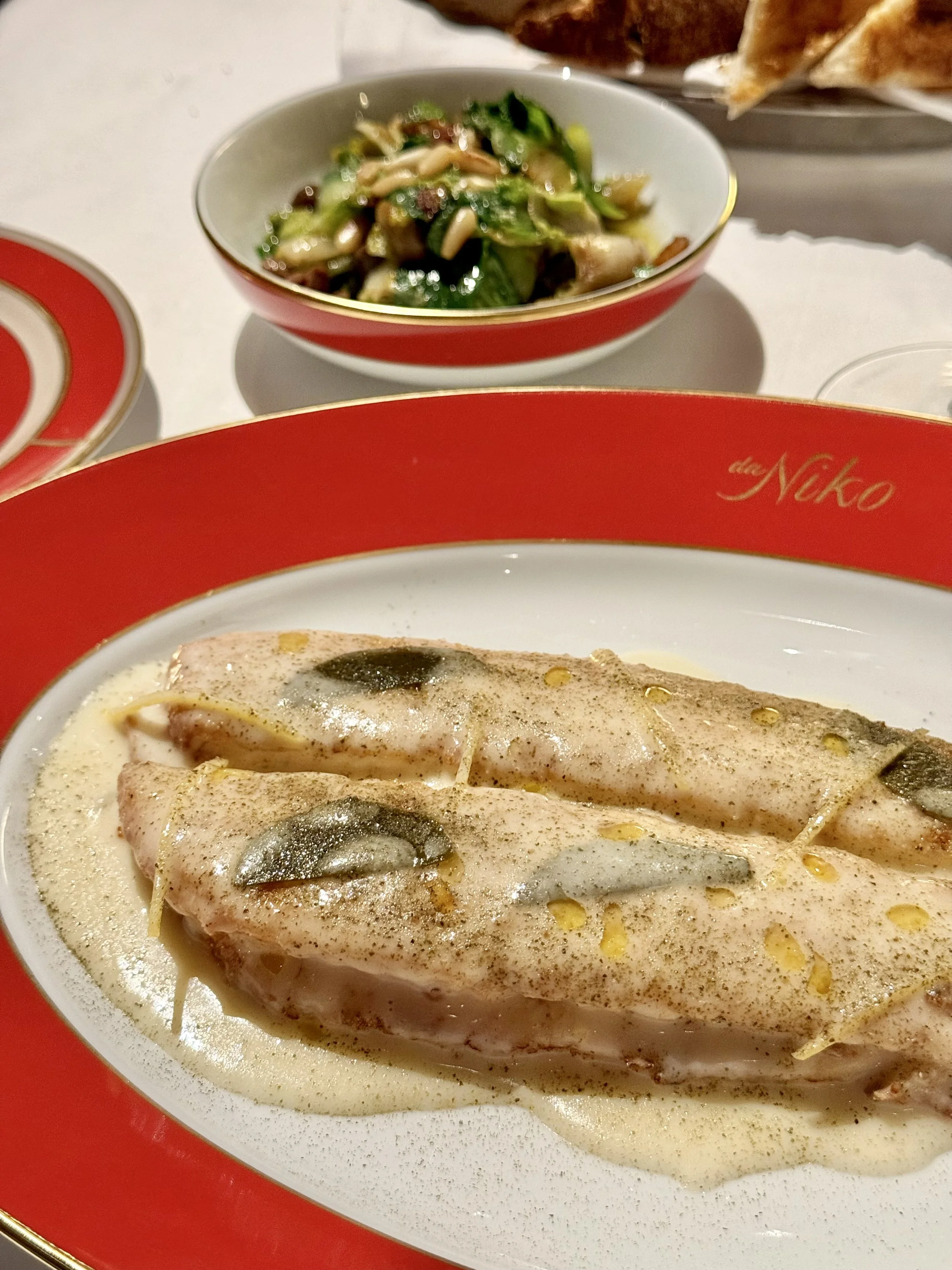 Sole fillets in a pale foamy sauce with green leaves and lemon strips, with a bowl of escarole and pine nuts behind.