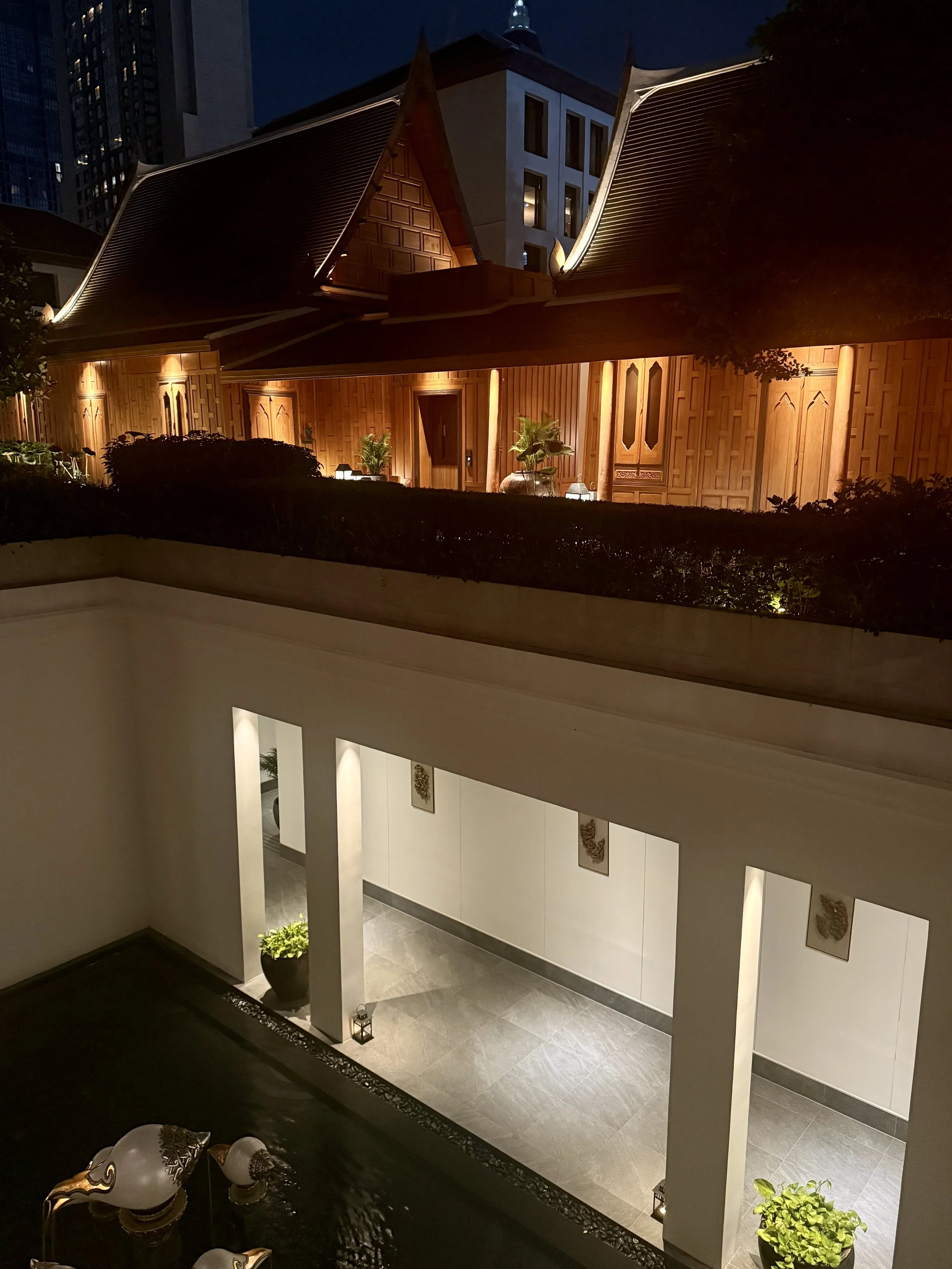 A warmly lit timber structure with a pitched roof stands above a white lower level at The Sukhothai Bangkok.