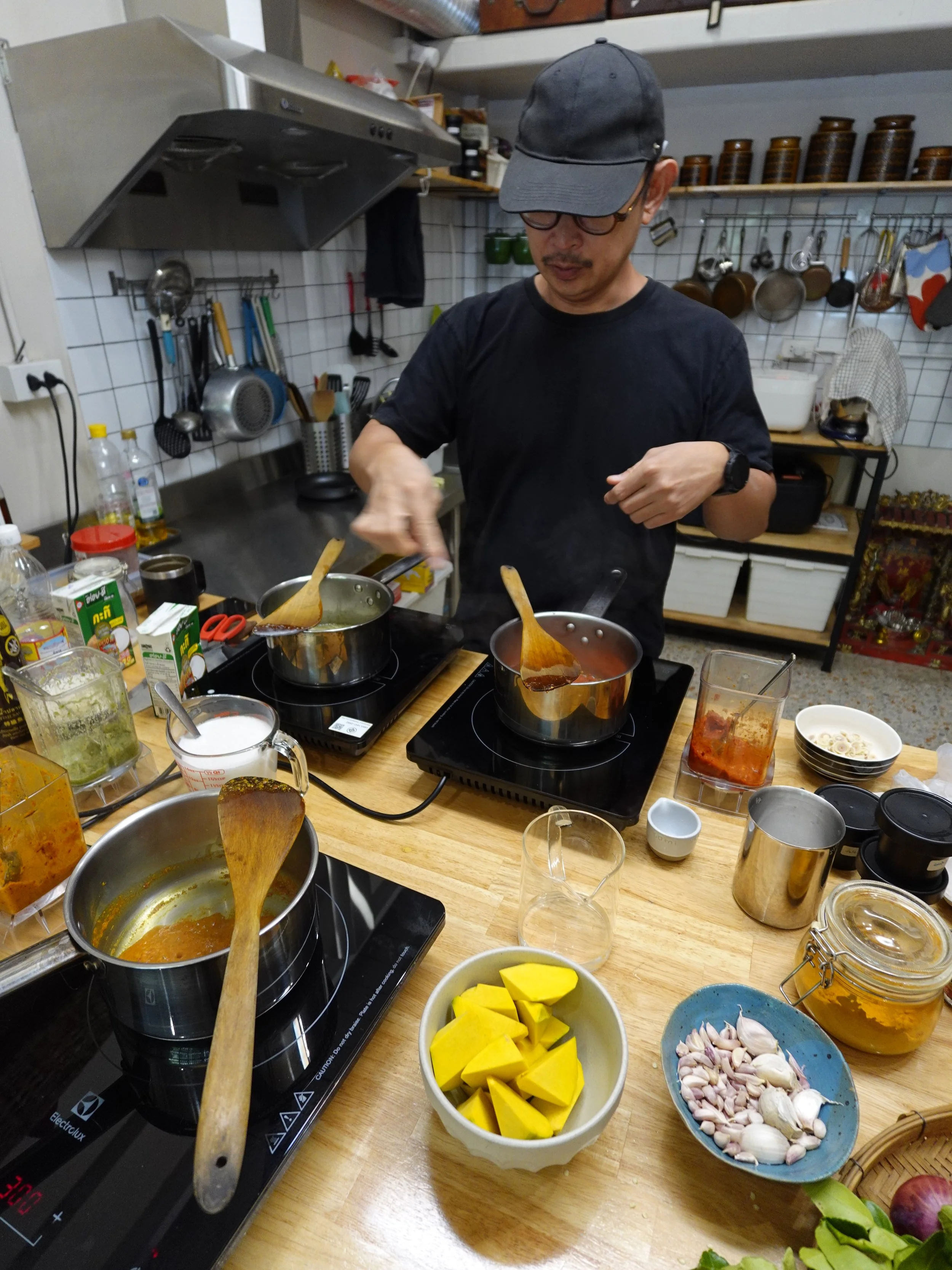 Angkrit at a wooden kitchen counter with three saucepans of curry, blender jugs, sliced pumpkin and a blue bowl of garlic.