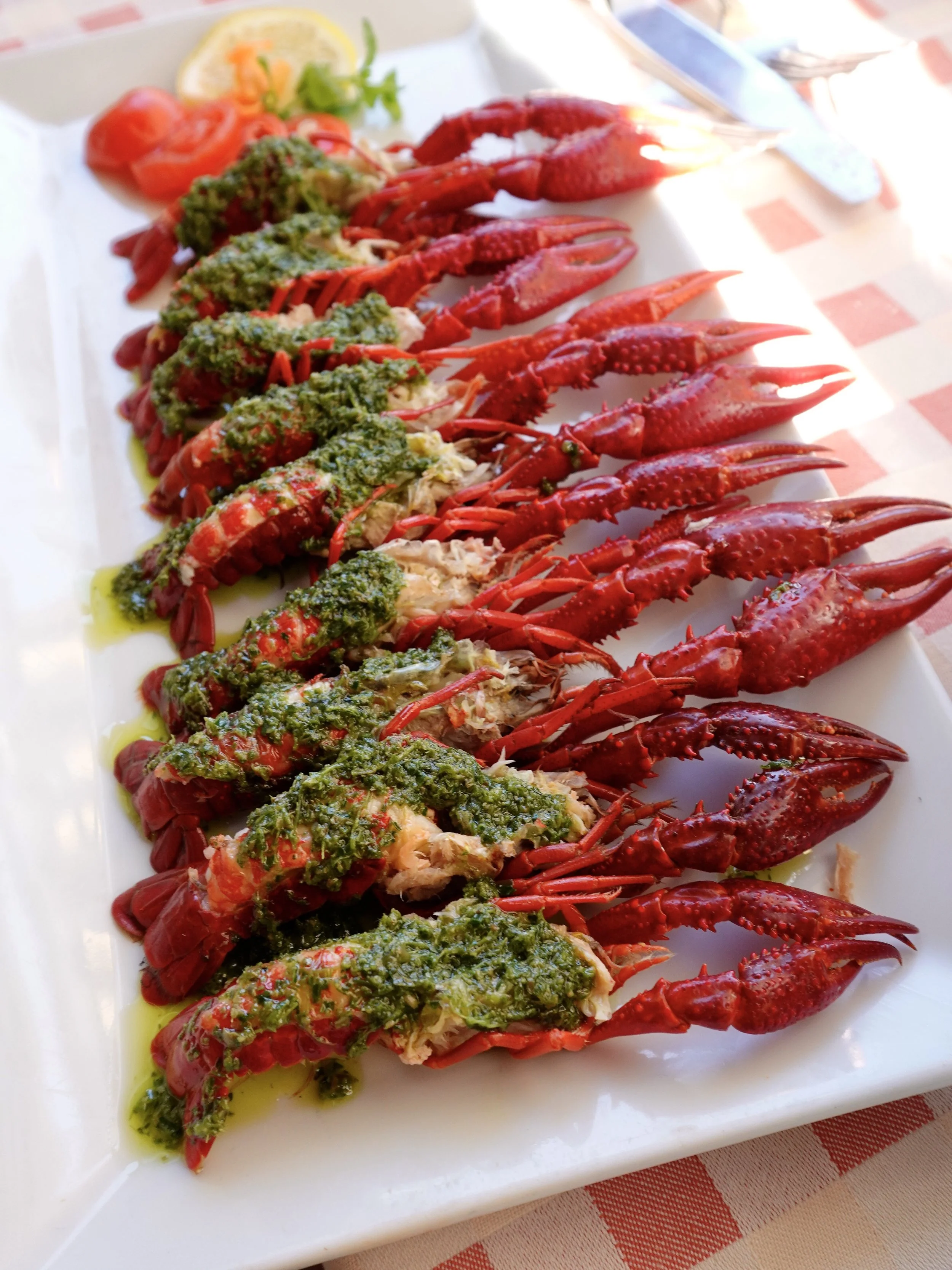A large white platter filled with vivid red freshwater crayfish, split open and covered in green salsa verde, on the checked tablecloth.