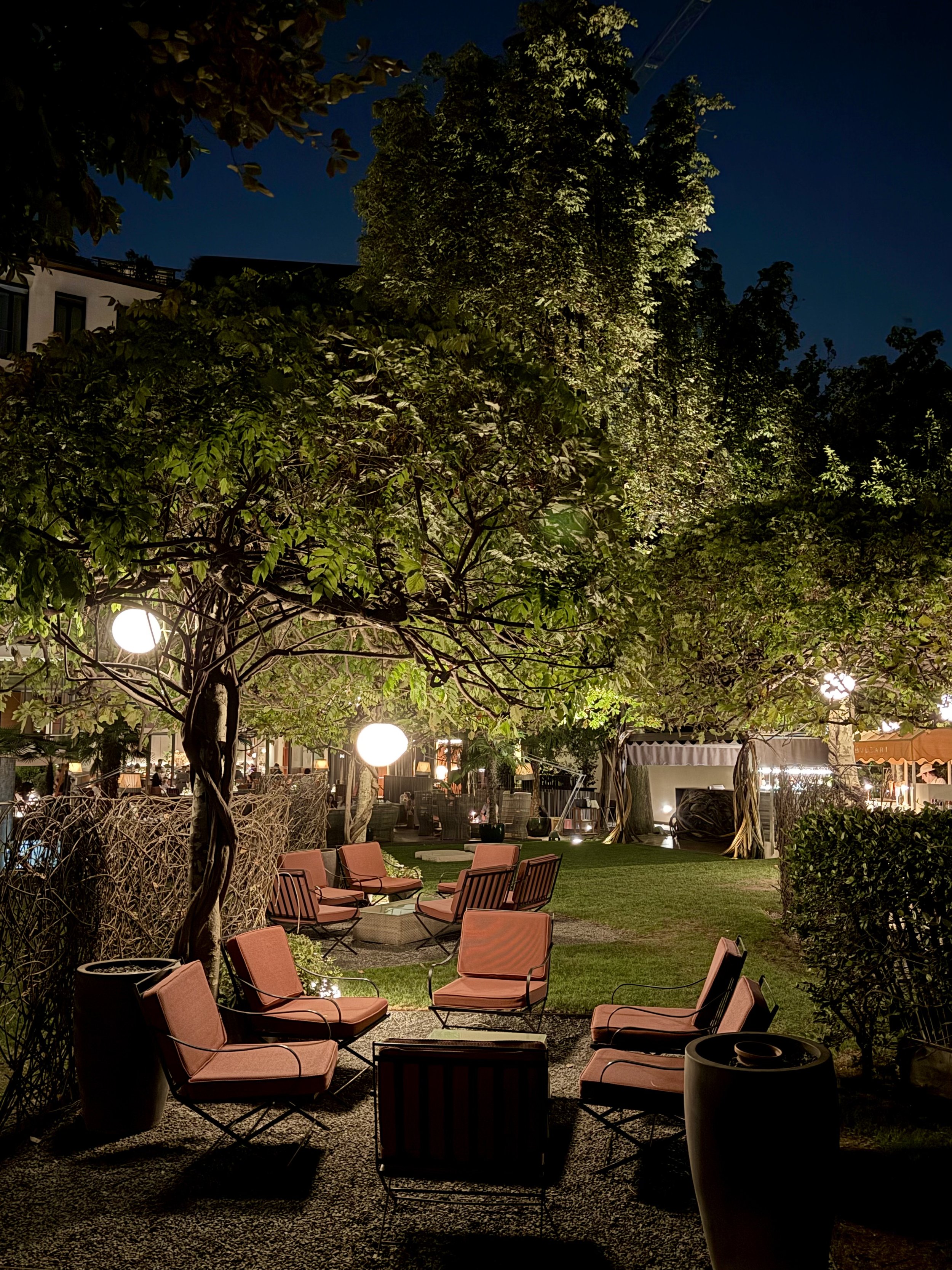 Bulgari Hotel Milano garden at night, with terracotta-coloured lounge chairs, low tables and globe lights among leafy trees.