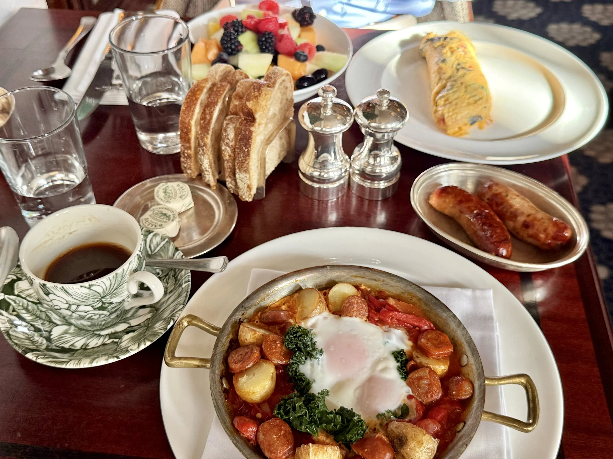 Breakfast at Millie’s Lounge with baked eggs, chorizo, potatoes, peppers and kale, alongside an omelette, sausages, mixed fruit, toast and coffee in a green-patterned cup.